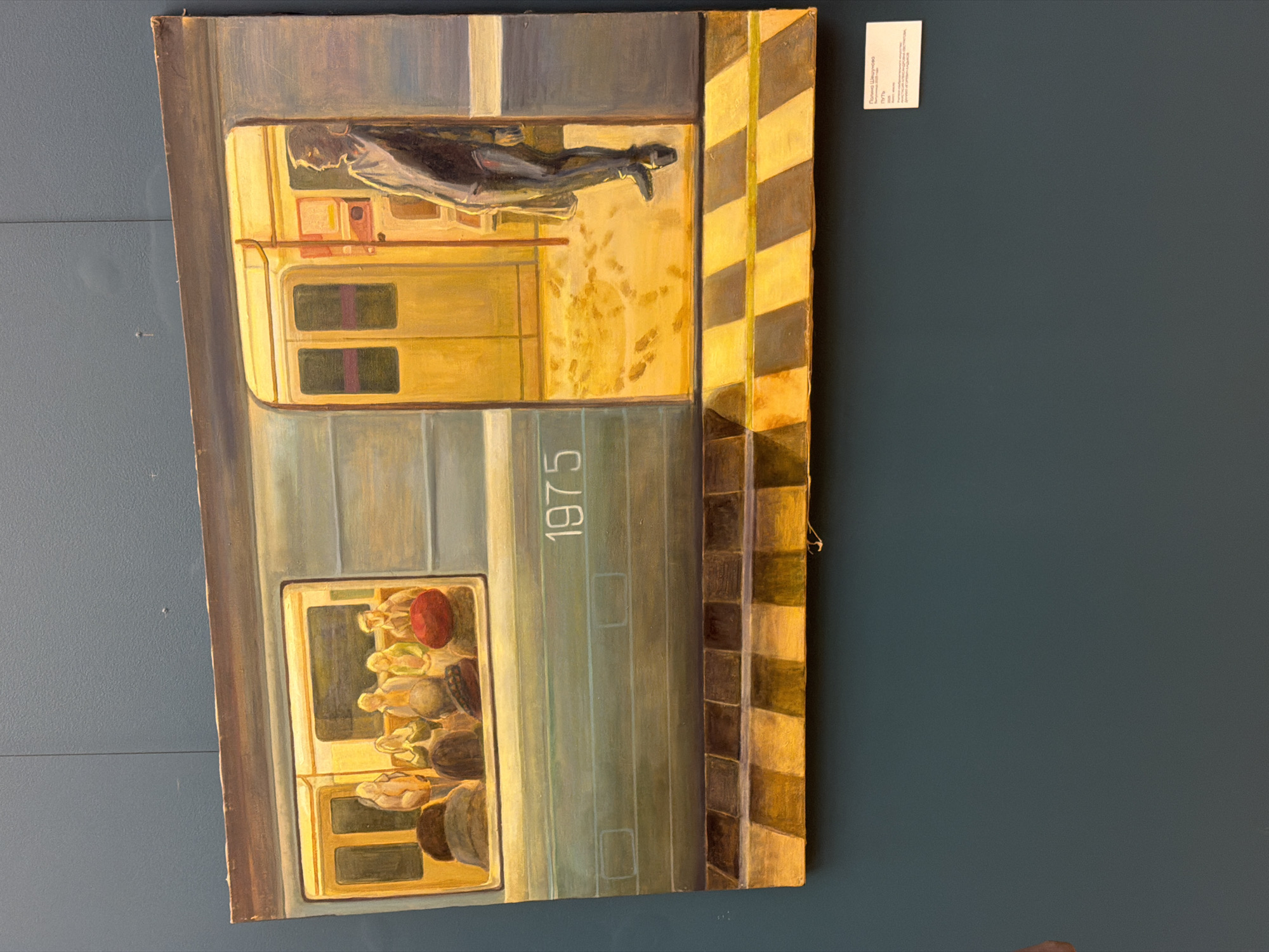 The image is a painting of a Moscow Metro train car from 1975. The painting depicts two scenes. One scene shows a man lying across the seats in the upper section of the train. The second scene shows several passengers sitting in the lower section of the train. The train car is painted with the year 1975. The side of the train car is next to a striped wall. The painting is hung on a blue wall.
