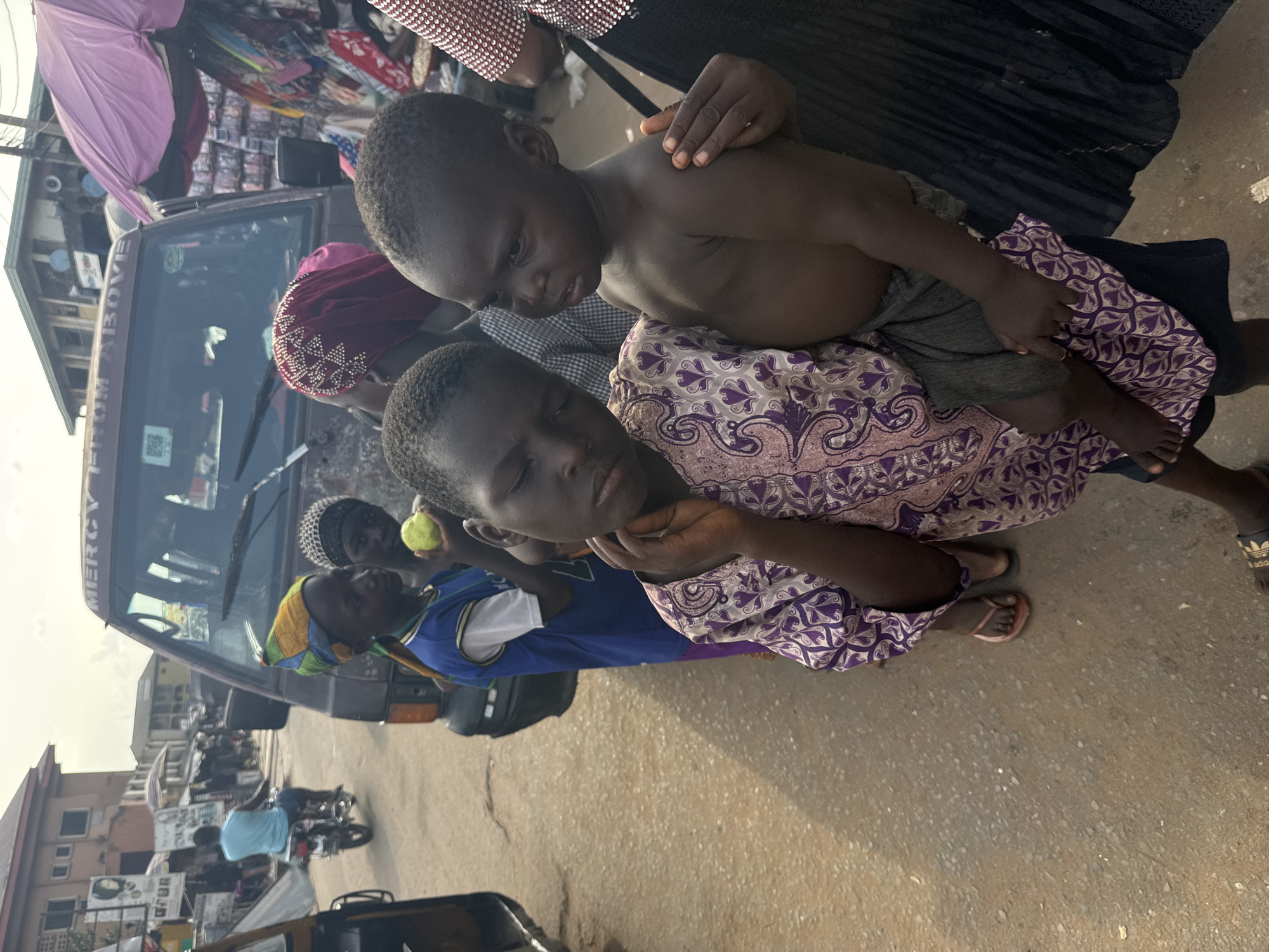 The image, taken in Onitsha, Nigeria, depicts a busy street scene, likely an outdoor market, during daylight hours under sunny conditions.
In the foreground, a woman in a purple and white patterned dress holds a young, shirtless child wearing grey shorts. The child looks forward, while the woman's head is tilted back with her eyes cast downwards, her left hand near her neck. She wears pink flip-flops.
Behind her, a child in a blue and white top and a colorful striped headscarf is visible, holding a light green fruit. Another child with a black and white checkered head covering is partially seen further back. A brown van or minibus dominates the midground, with the word "MERCY" and part of a "T" visible on its side above the windshield, which also displays a QR code sticker. Another individual, wearing a maroon head covering with white embellishments, is visible near the van's window.
To the upper left, a partially open purple umbrella suggests a market stall. The background features buildings lining the street, with a person on a motorcycle in the distance. The ground is paved with light-colored concrete or asphalt. The foot of another person, wearing a black Adidas slide, is visible on the right.