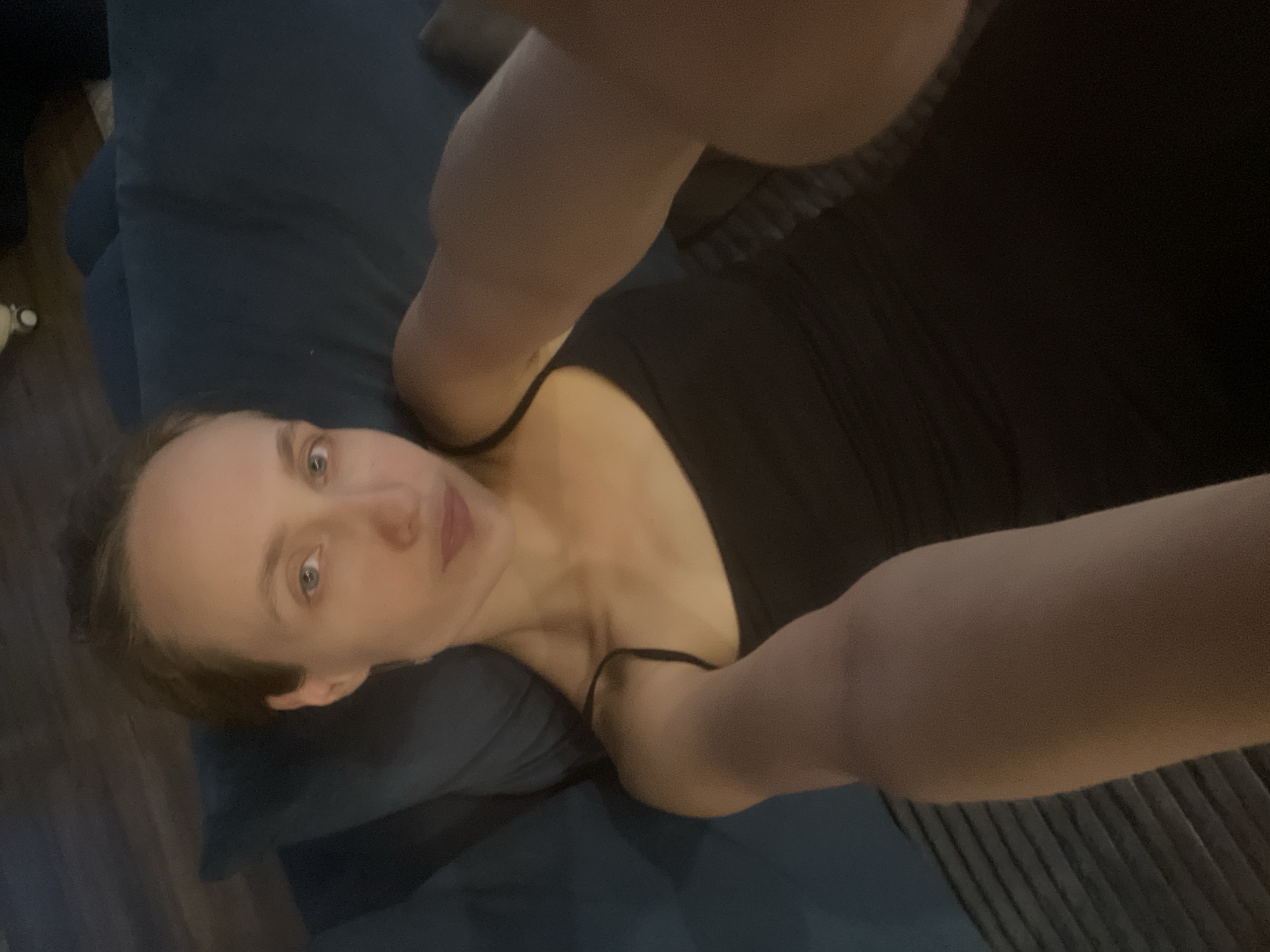 A person is lying on their side on a dark blue surface, likely a couch or bed. They are wearing a black tank top. Their right arm is extended, holding what appears to be a phone, taking a selfie. The person's face is visible, looking towards the camera with a neutral expression. Their eyes are open and looking directly at the viewer. The background consists of the dark blue surface and some wooden flooring. The lighting is dim, suggesting it might be nighttime or indoors with low light. There are no visible signs of specific weather conditions. The setting appears to be a casual, indoor environment.