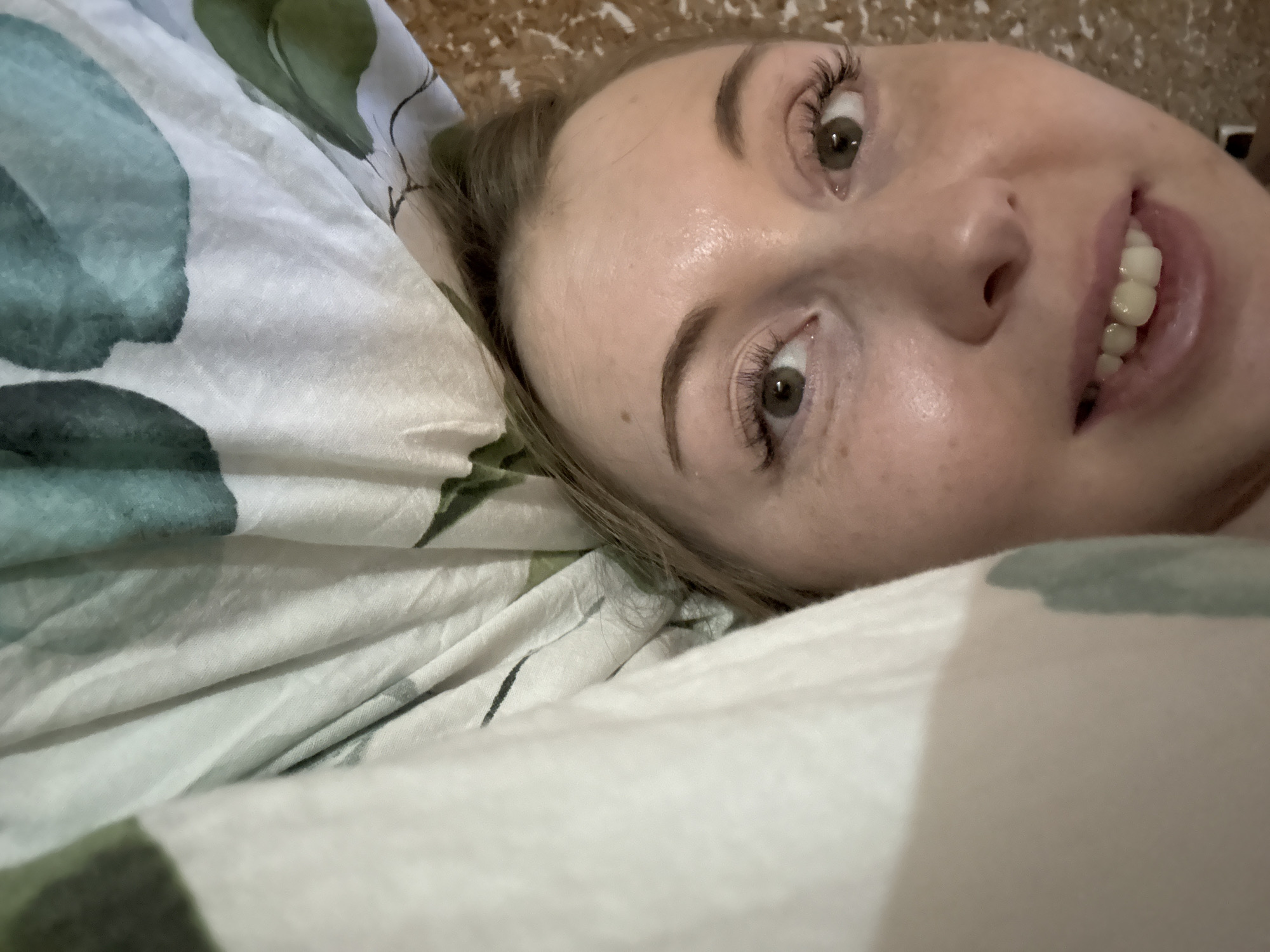 A close-up shot focuses on a woman with fair skin, lying in bed. Her face is the central subject, displaying light freckling and brown eyes. Her lips are slightly parted, revealing her teeth. She has brown eyebrows and prominent eyelashes.
The woman appears to be lying on a bed with patterned sheets. The sheets feature a design of white fabric with teal and green floral patterns.
The background is blurred and out of focus. The location appears to be indoors, within a bedroom setting.