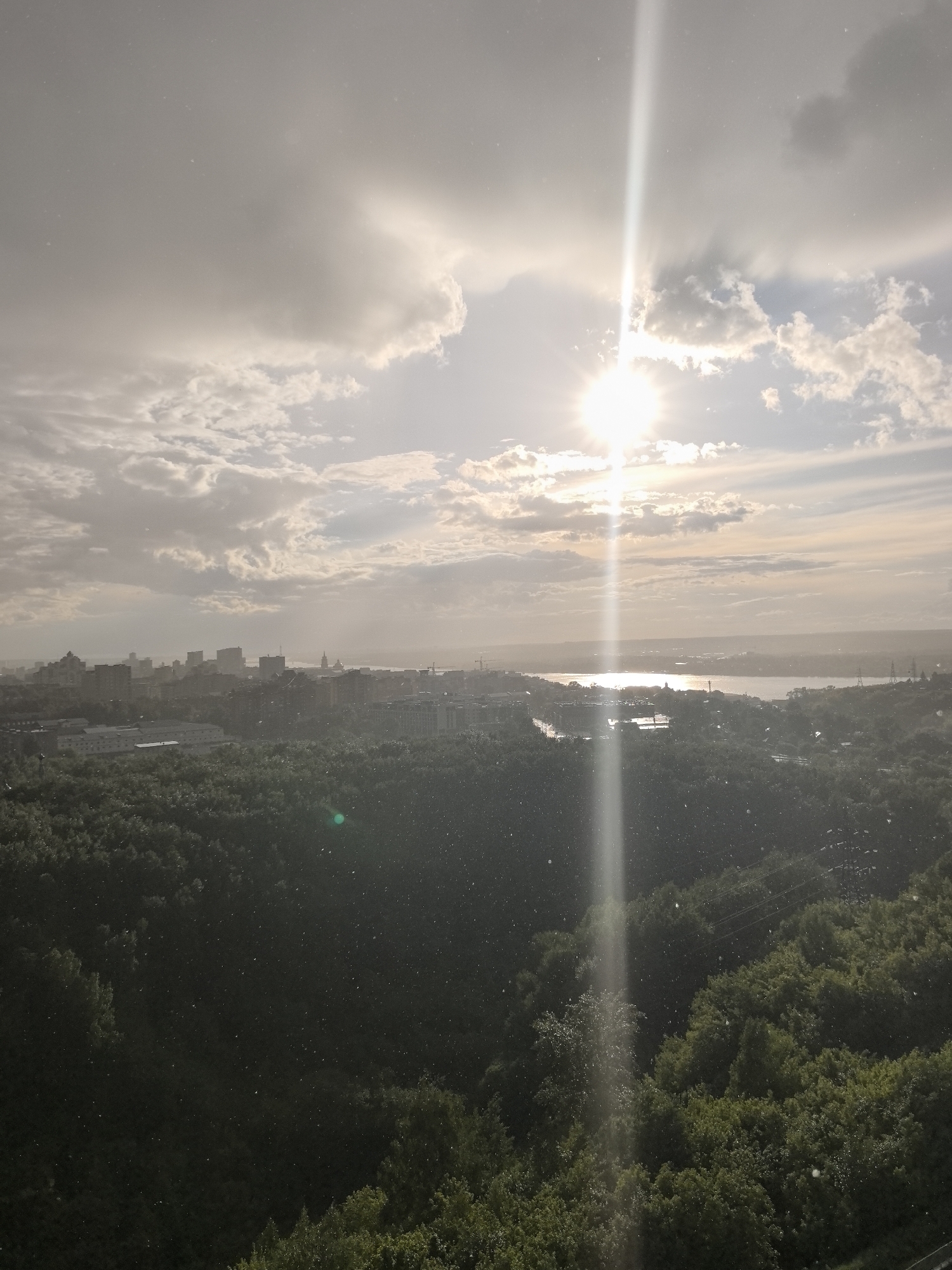The image shows an aerial view of Perm, Russia, captured during a rain shower with the sun shining through the clouds. The city skyline is visible in the distance, with buildings and structures discernible but obscured by the weather. A body of water, likely the Kama River, reflects sunlight. Dense forest covers the foreground. The sun is positioned centrally in the sky, creating a vertical glare in the image. Raindrops or particles are visible throughout the scene.