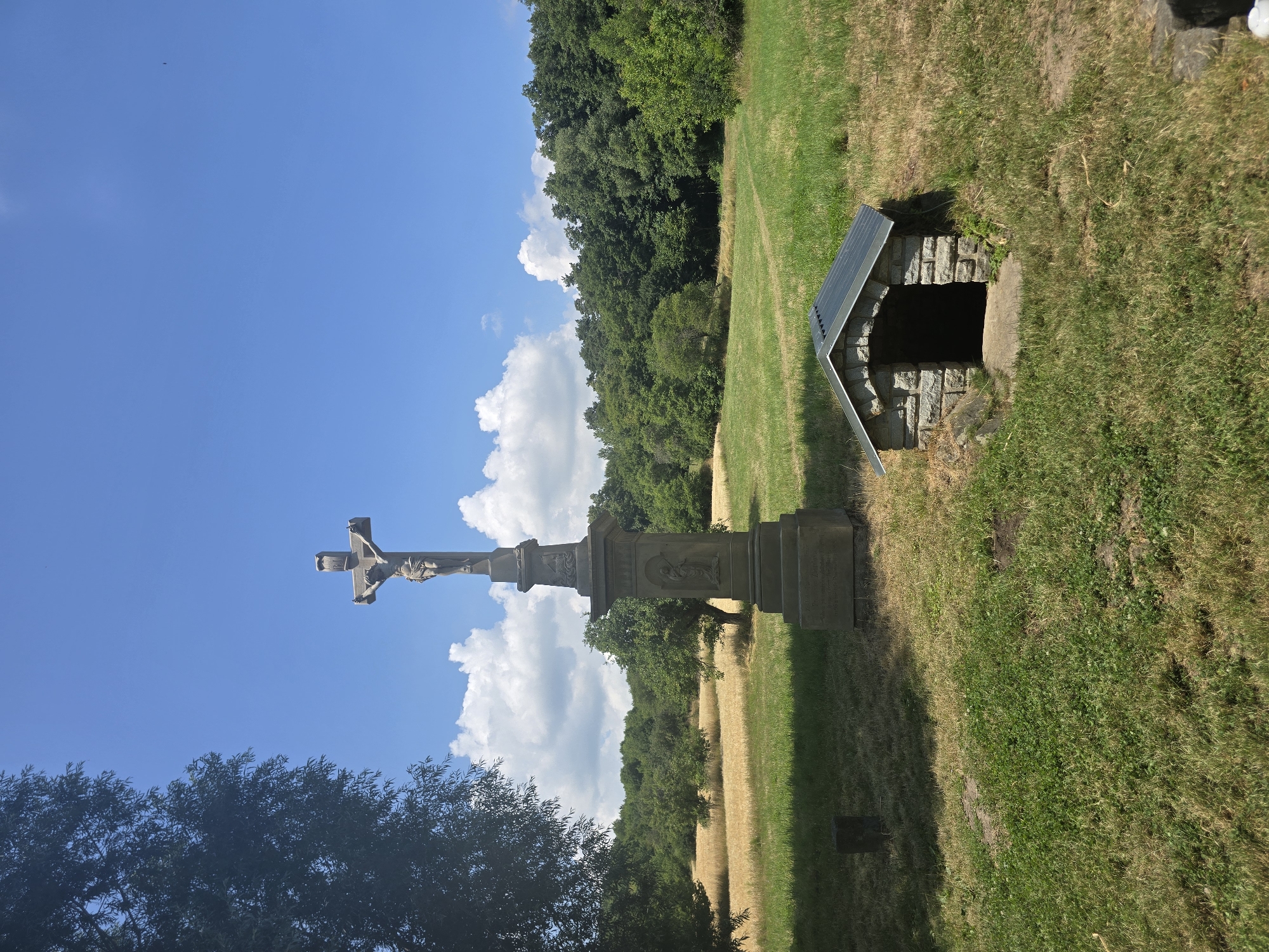 In Pustiměř, Czechia, on a sunny day with a partly cloudy sky, a stone crucifixion monument stands tall in a grassy field. The monument features a detailed sculpture of Jesus Christ on the cross, carved from stone. To the right of the monument is a small, arched stone structure with a metal roof, possibly a well or a small shelter. The field is bordered by a dense forest in the background and a field of yellow crops on the left. The overall scene conveys a sense of historical and religious significance within a natural landscape.