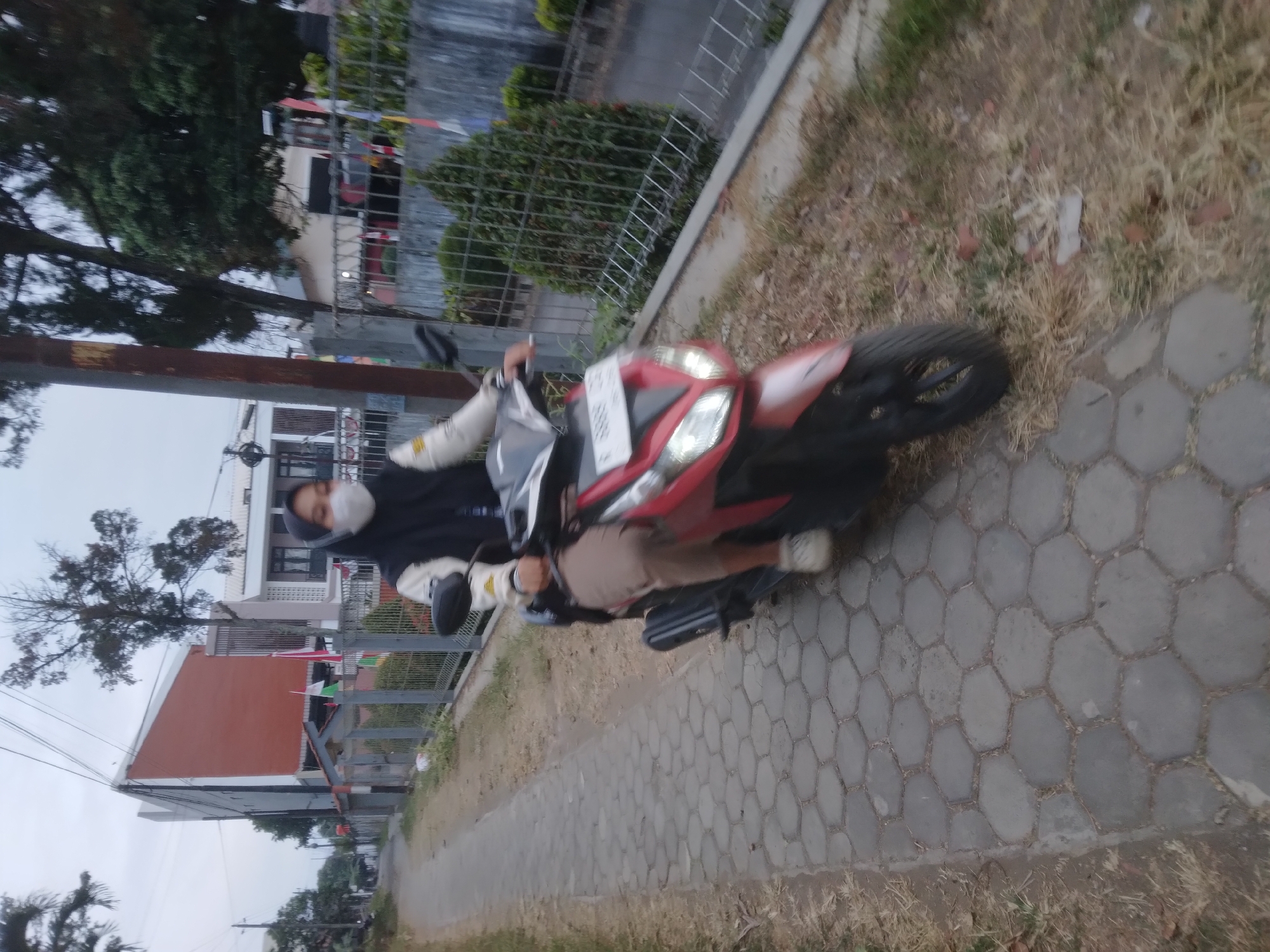 A medium shot captures a person riding a red and black scooter on a paved pathway in a residential area of Desa Pamijen, Kecamatan Sokaraja, Indonesia. The person, wearing a black hijab, a light-colored jacket with black accents, and tan pants, is the central subject. They are also wearing a white face mask. The scooter has a license plate with the number "B 5888 CJG". The pathway is made of hexagonal grey paving stones and is bordered by dry grass and foliage on the right. In the background, to the left, is a modern, multi-story building with an orange facade and white trim, surrounded by a fence. To the right of the path, a metal fence encloses a yard with manicured green bushes and trees. The sky is overcast and the overall lighting suggests late afternoon or early morning. The image appears to have been taken from a slightly elevated position.