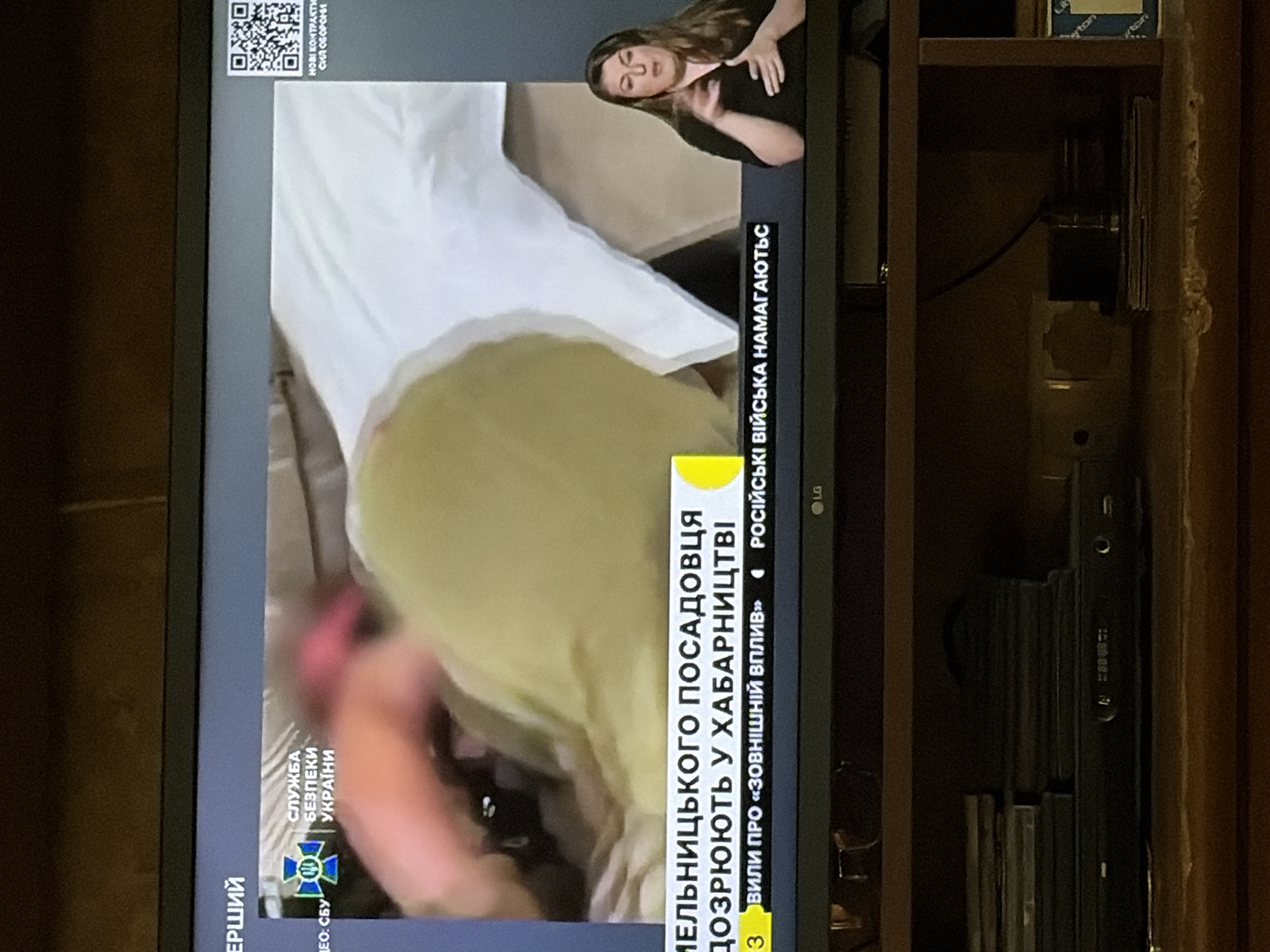 The image shows a television screen displaying a news broadcast. In the foreground, a woman is signing on the right side of the screen. The news broadcast itself shows a graphic with the text "СЛУЖБА БЕЗПЕКИ УКРАЇНИ" (Security Service of Ukraine) on the left. Below this, text in Ukrainian reads "МЕЛЬНИЦЬКОГО ПОСАДОВЦЯ ДОЗРЮЮТЬ У ХАБАРНИЦТВІ" which translates to "Melnytsky's official is suspected of bribery". Further text reads "ВИЛИ ПРО «ЗОВНІШНІЙ ВПЛИВ» РОСІЙСЬКА ВІЙСЬКА НАМАГАЮТЬСЯ" which translates to "about 'external influence' Russian troops are trying". A QR code is visible on the top right of the screen. The television is mounted above a shelf containing electronic devices and media.