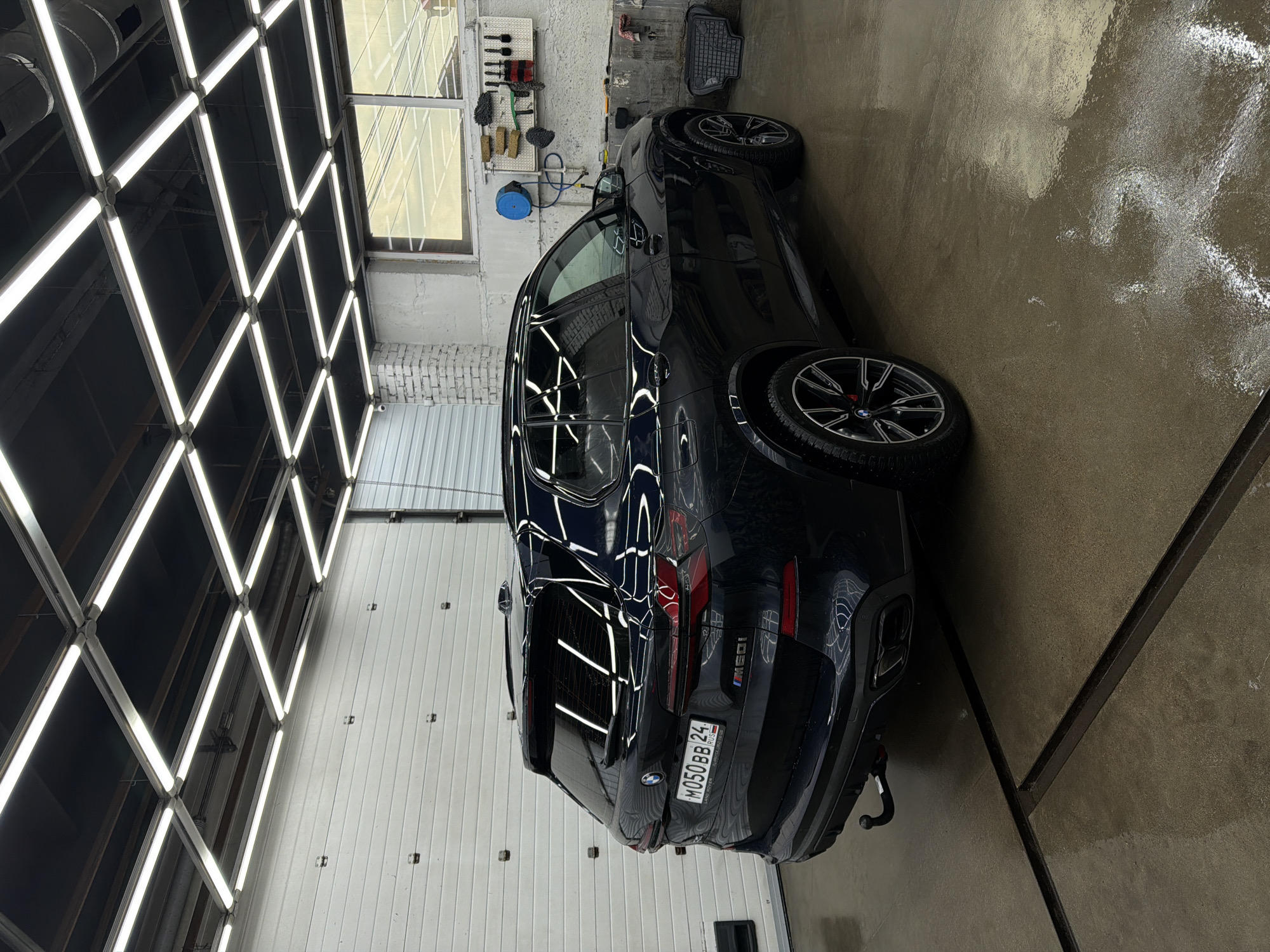 The image depicts a black BMW X5 M50i parked indoors, likely in a detailing or car wash facility. The car's rear is visible, showcasing its M badge, rear lights, and license plate "M050BB24" indicating registration in Krasnoyarsk, Russia (Region code 24). The floor is wet, suggesting recent cleaning or washing. The environment includes a garage-like space with a grid of fluorescent lights on the ceiling and a white wall. There's cleaning equipment mounted on the wall, implying a professional car cleaning setting. There are no people visible in the scene.