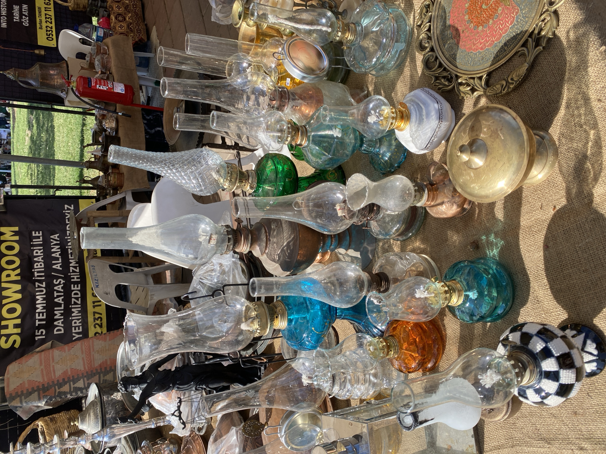 The image depicts a collection of vintage or antique items displayed on a burlap-covered surface. The primary objects are numerous glass oil lamps in various shapes, colors, and styles, with some lamps having ornate metal bases. Adjacent to the lamps, there's a vintage-looking framed ornamental plate. In the background, one can discern a fire extinguisher, white plastic chairs, and a sign for "SHOWROOM" located in Damlataş/Alanya, Turkey, with contact information. The setting appears to be an outdoor market or vendor stall, possibly related to antiques or historical artifacts. There are no people visible in the main focus of the image.