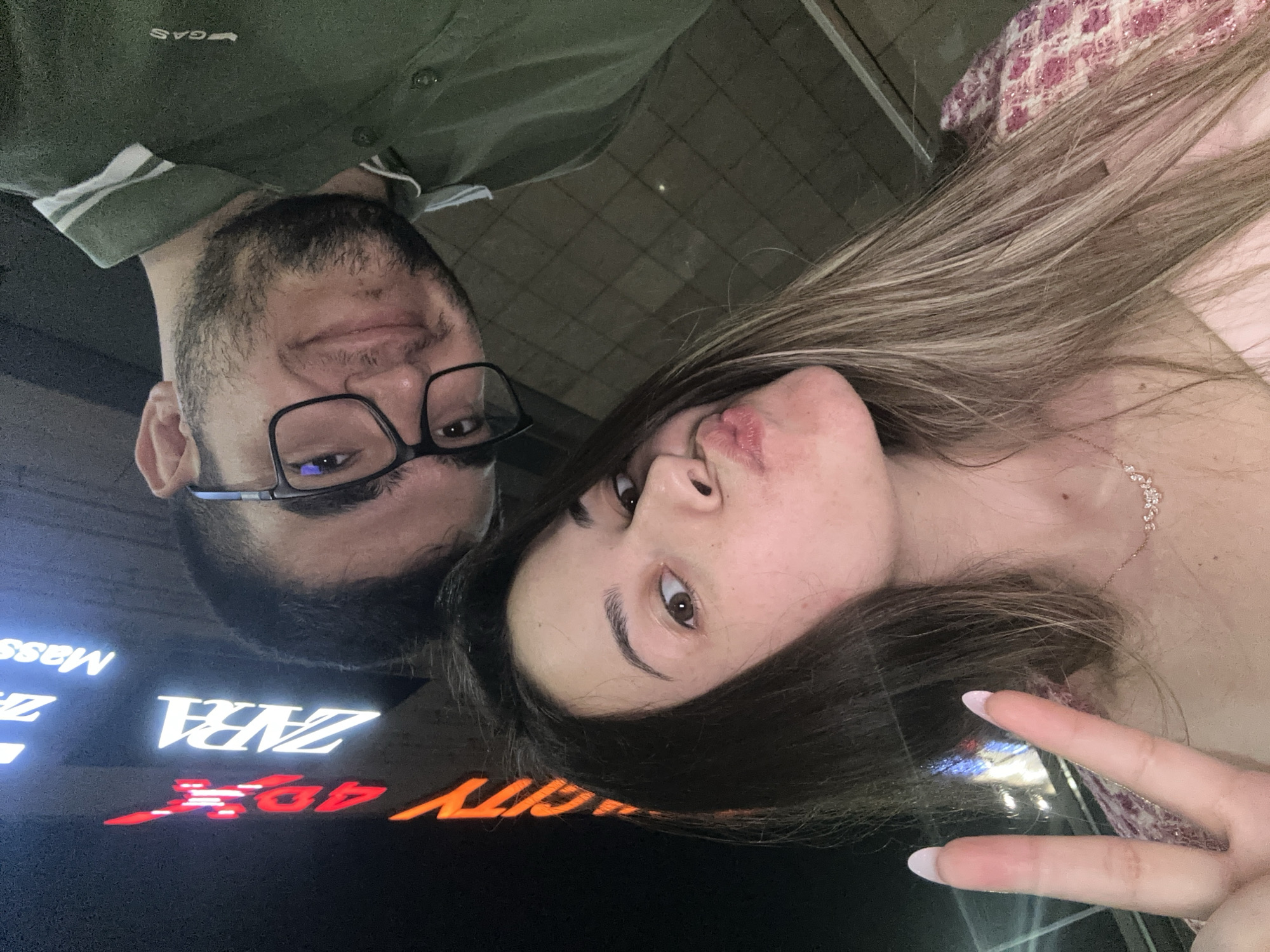 A selfie-style photograph captures a young adult couple in an urban setting at night. In the foreground, a young adult female with long, dark hair featuring lighter streaks looks directly at the camera with pouted lips. She wears a delicate necklace, a light-colored top, and a pink and white patterned jacket. Her left hand is raised, making a peace sign, displaying long, manicured nails with light polish. Behind her and slightly above is a young adult male with short dark hair, a beard, and black-framed glasses. He looks towards the camera with a neutral expression, wearing a dark green collared shirt with two white stripes on the placket and a "GAS" brand logo on the shoulder. The background is dark, illuminated by various commercial neon signs, including "ZARA" in white, a partial red "Adidas" logo, and "CITY 4DX" in orange/red, alongside other indistinct white text, likely "Mass" or "Massi." This combination of brands strongly suggests the location is within a major shopping mall in Sofia, Bulgaria, such as Paradise Center, The Mall, or Park Center Sofia (formerly City Center Sofia), which are known to host both ZARA stores and Cinema City 4DX theaters.