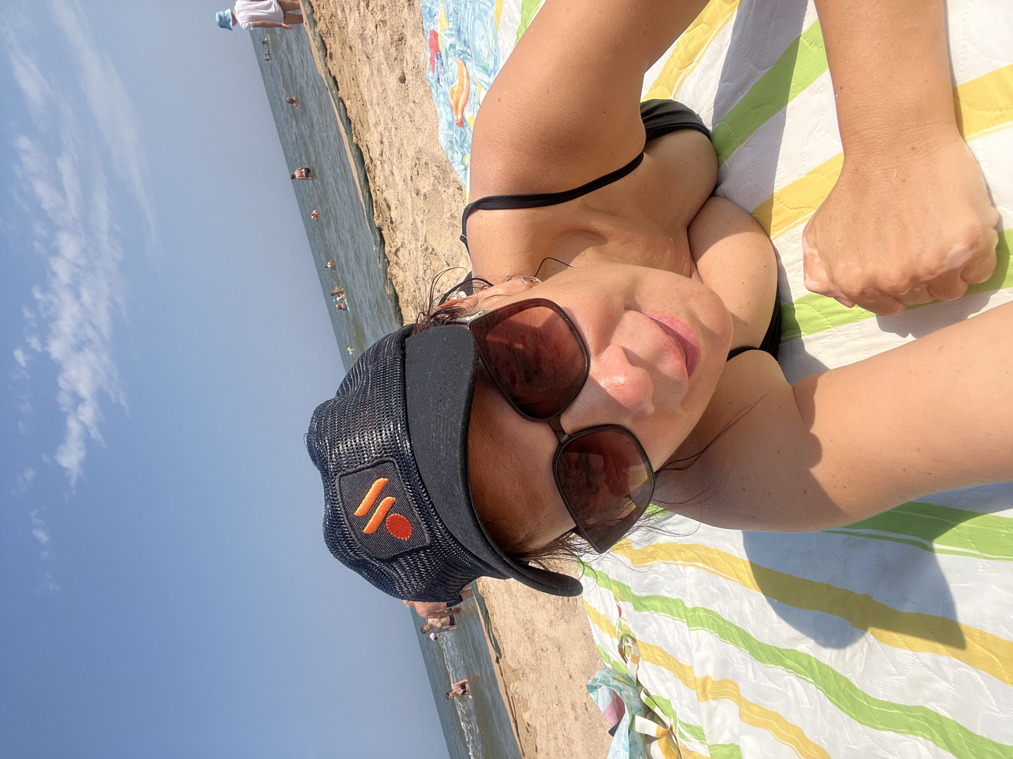A woman lies on a colorful striped beach towel on a sandy beach, facing the camera in a selfie. She is wearing a black bikini top, a black baseball cap with an orange logo, and large sunglasses. In the background, the sea is visible with several people swimming and wading in the water. The sky is clear blue with a few scattered clouds. The scene appears to be a sunny day at the beach in Golubitskaya, Russia, a resort town on the Taman Peninsula known for its sandy beaches and shallow waters.