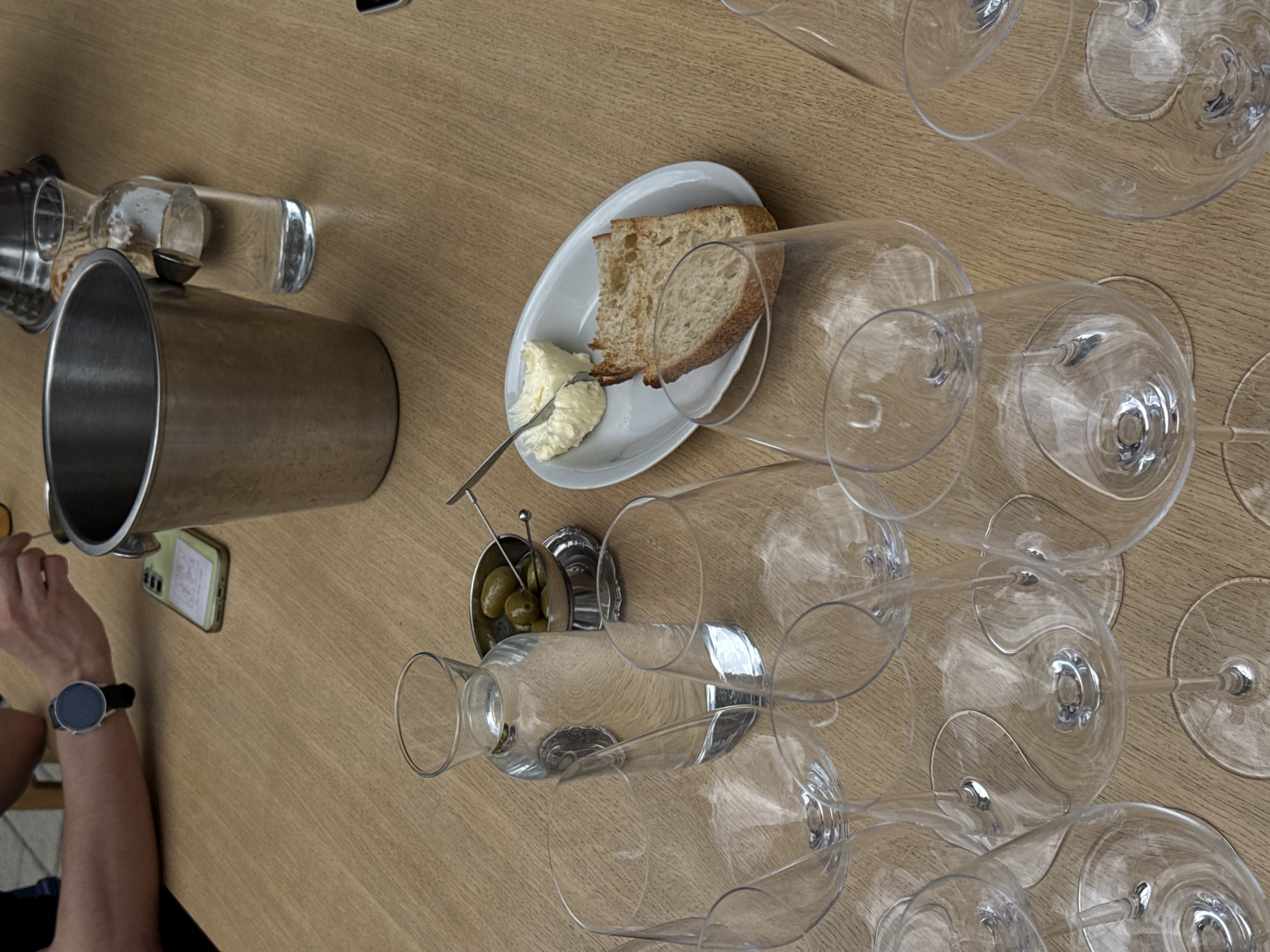 The image shows a table setting, likely in a restaurant in Belgrade, Serbia. There are multiple empty wine glasses arranged across the table, along with a white plate containing bread and butter, and a small metal container of olives. A metal ice bucket sits near the edge of the table, next to a phone. A person's arm and wrist are visible, with a watch on their wrist, seemingly reaching for something with a small utensil. The table has a light brown wood grain finish.