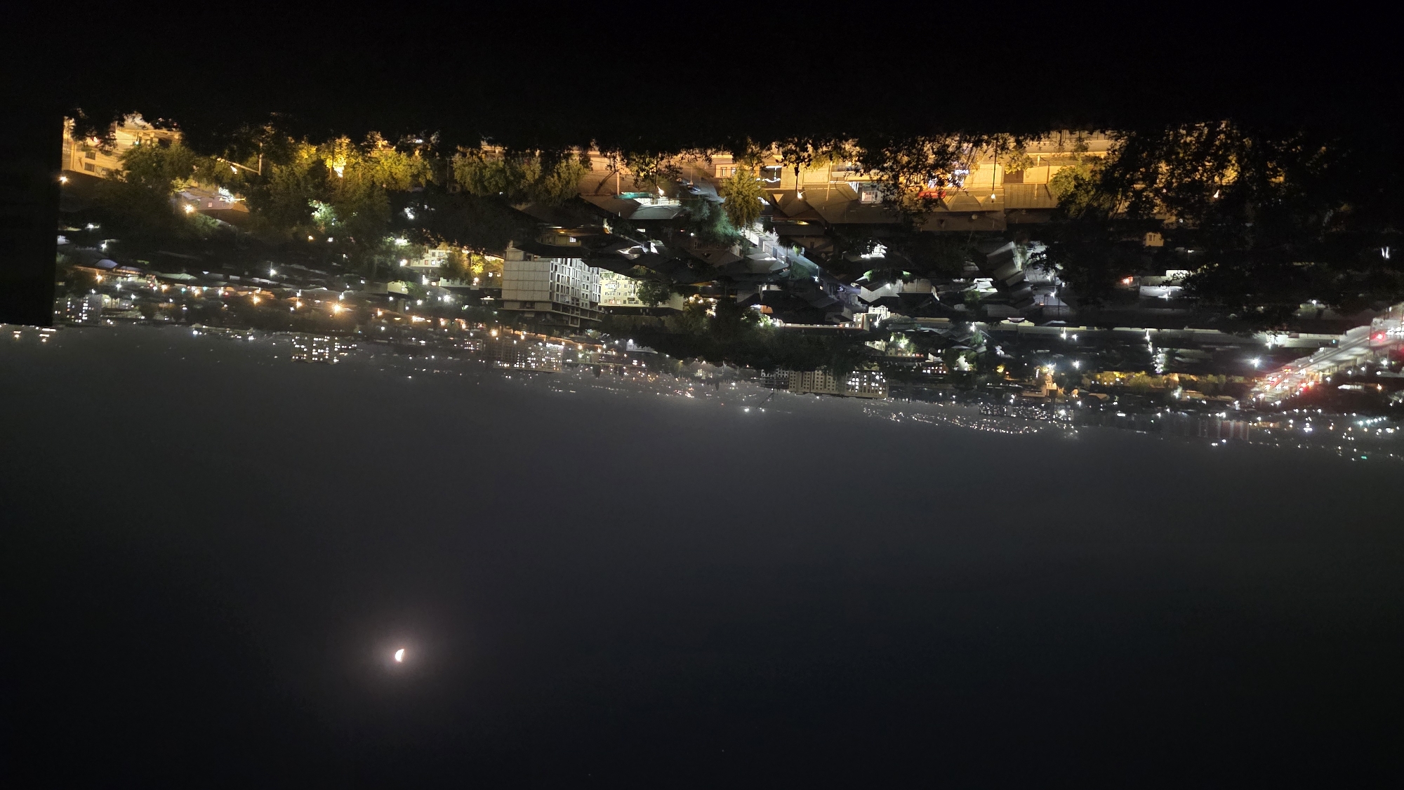 This is a nighttime, high-angle, wide shot of the city of Osh, Kyrgyzstan. The image captures a sprawling urban landscape illuminated by streetlights and building lights. A crescent moon is visible in the upper right quadrant of the dark sky. The foreground is dominated by silhouetted trees and foliage, obscuring a portion of the view. Below the trees, a dense residential area with numerous houses and some multi-story buildings is visible. Streets are lined with lights, suggesting ongoing traffic. In the distance, the cityscape extends towards a hazy horizon, with more lights indicating further development and infrastructure. The overall atmosphere is one of a calm, illuminated city at night.