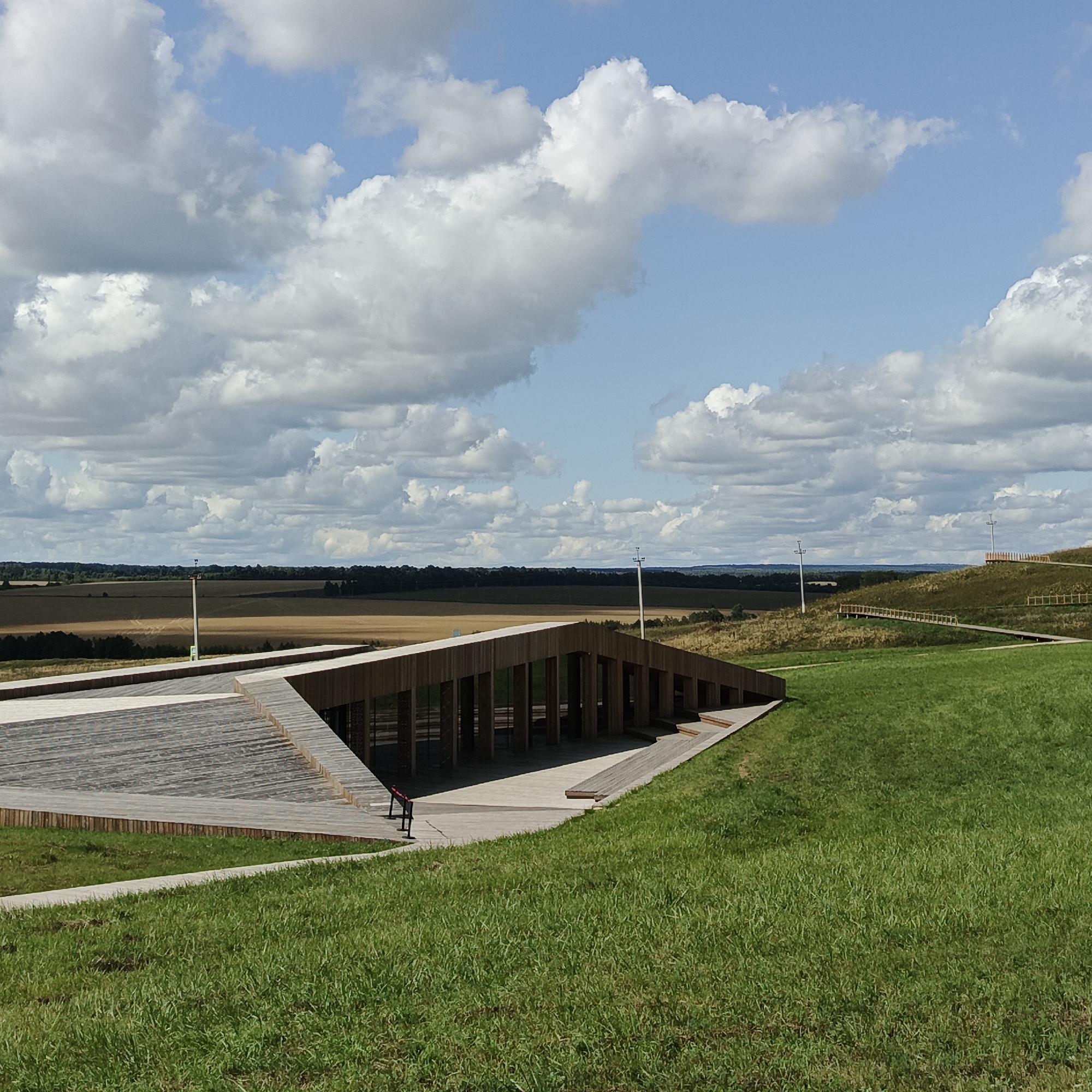 This is a full, eye-level shot of an outdoor park-like setting in Verkhnie Termy, Russia, on a clear day with fluffy white clouds against a blue sky. In the foreground, a modern wooden structure with a sloped roof and a large open-air seating area made of wooden planks is visible. The structure features vertical wooden pillars supporting the roof. Adjacent to the wooden structure is a well-maintained green lawn. In the mid-ground, rolling grassy hills stretch into the distance, dotted with patches of dry vegetation and trees. Several wooden boardwalks with railings wind up the hills. Two tall, white poles with lights or antennas are prominent on the landscape, one on a hill and another in the middle ground. A distant treeline forms a dark band across the horizon, beyond which lies a flat, cultivated field. The overall scene is serene and showcases a blend of natural landscape and architectural elements. No people are visible in the image.