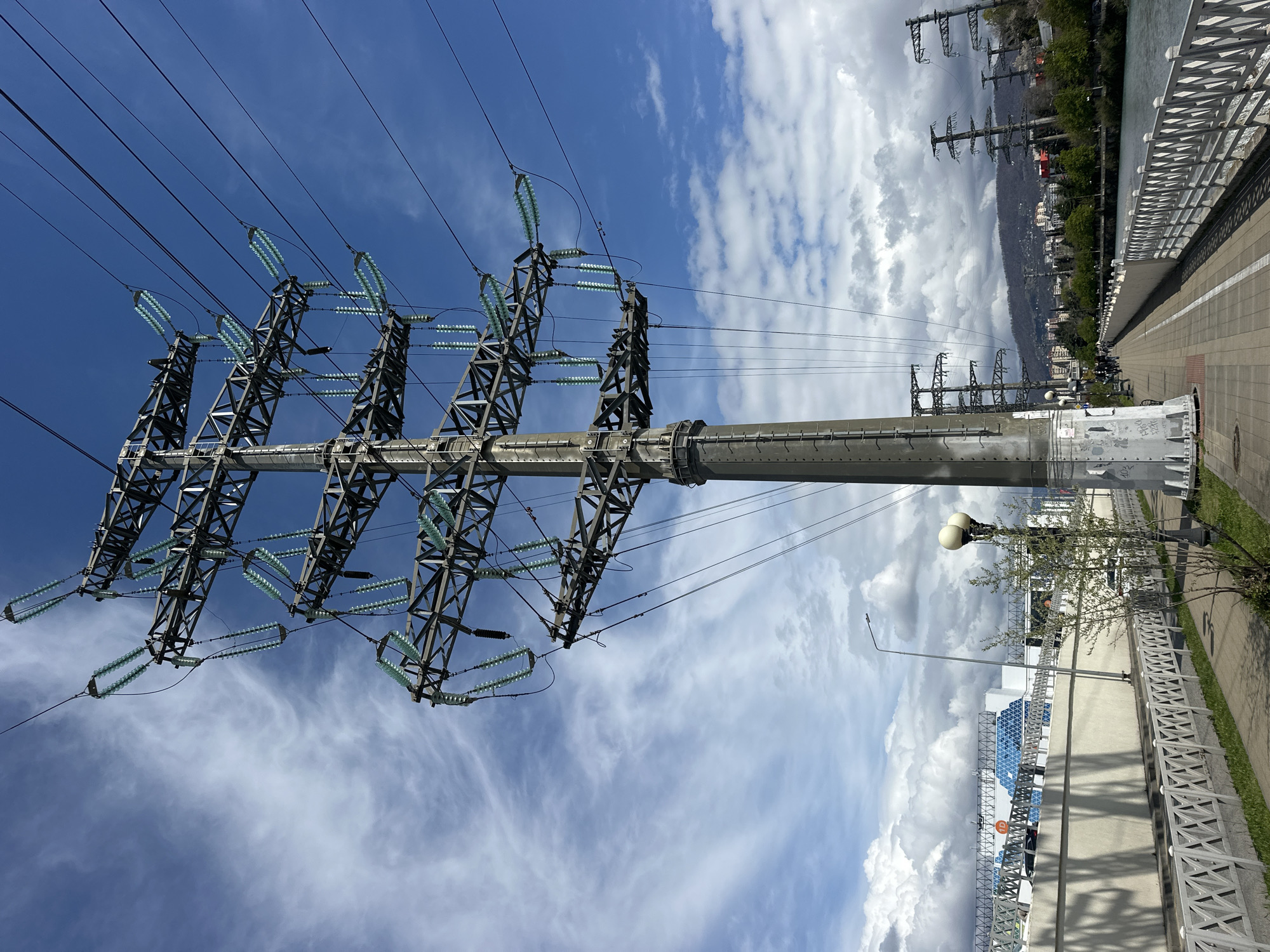 The image shows a large metal electrical power tower located on a concrete walkway in Sochi, Russia. The tower has a cylindrical base and a lattice-like structure at the top supporting multiple electrical insulators and power lines. The sky is visible behind the tower, filled with clouds. The walkway is bordered by a metal fence and green grass on the right. A light post and building can be seen in the background. In the distance, more electrical towers are visible, as well as buildings and foliage on a distant hillside.