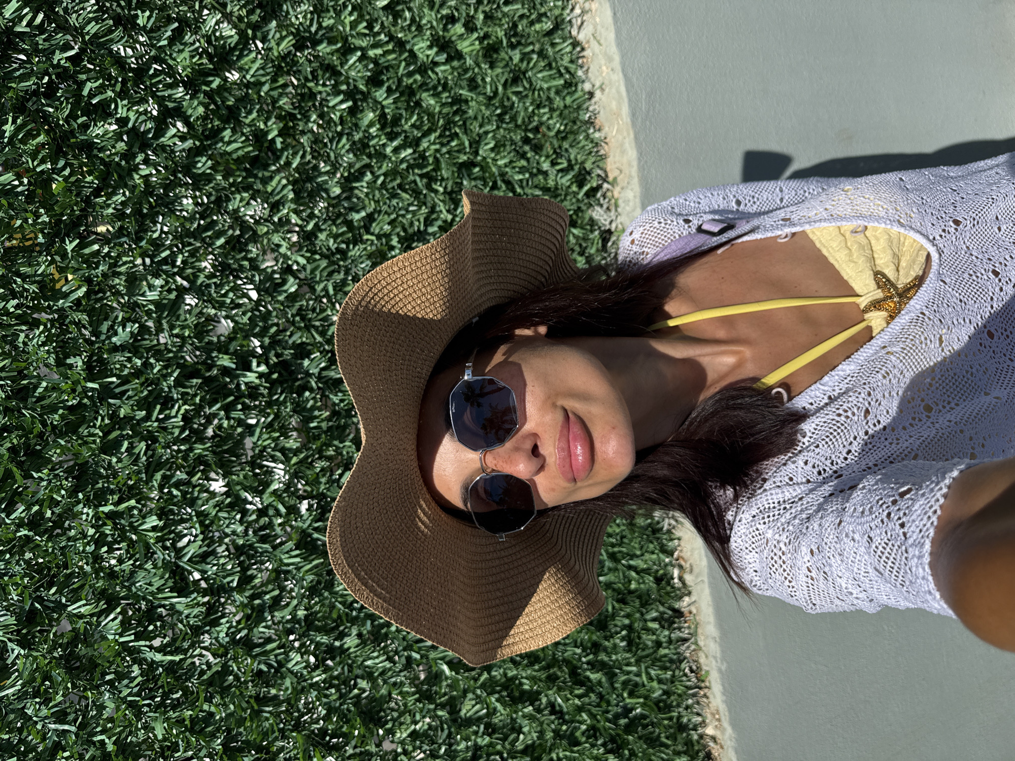 A woman wearing a straw hat and sunglasses poses for a selfie in front of a green hedge wall. She is also wearing a yellow bikini and a white open-knit cover-up. The background appears to be an outdoor setting, possibly a walkway or patio. The lighting suggests it is daytime and sunny.