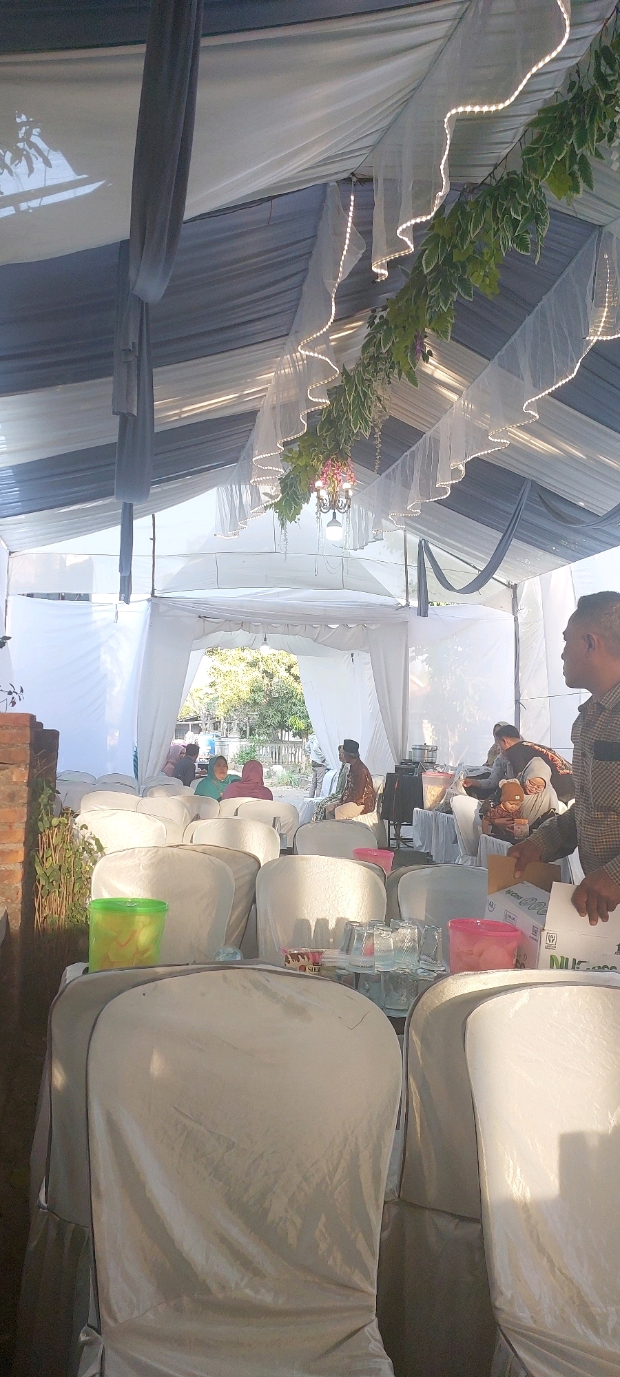 The image depicts an outdoor gathering, possibly a wedding reception, taking place under a white tent decorated with flowing grey and white fabric, fairy lights, and greenery. In the foreground, several white-covered chairs are visible, some with a dull sheen indicating a plastic or vinyl material. To the left, a green plastic container with a yellow lid sits on a surface. In the middle ground, through an opening in the tent, a group of people are seated and standing, dressed in various attire, including a man wearing a traditional peci hat. Further back, trees and a building can be seen, suggesting a rural or semi-rural setting. On the right side of the image, a man in a checkered shirt is holding a cardboard box. The lighting suggests it is daytime, with sunlight filtering through the tent and casting shadows. This scene is described as being in the city of Bukur, Indonesia.