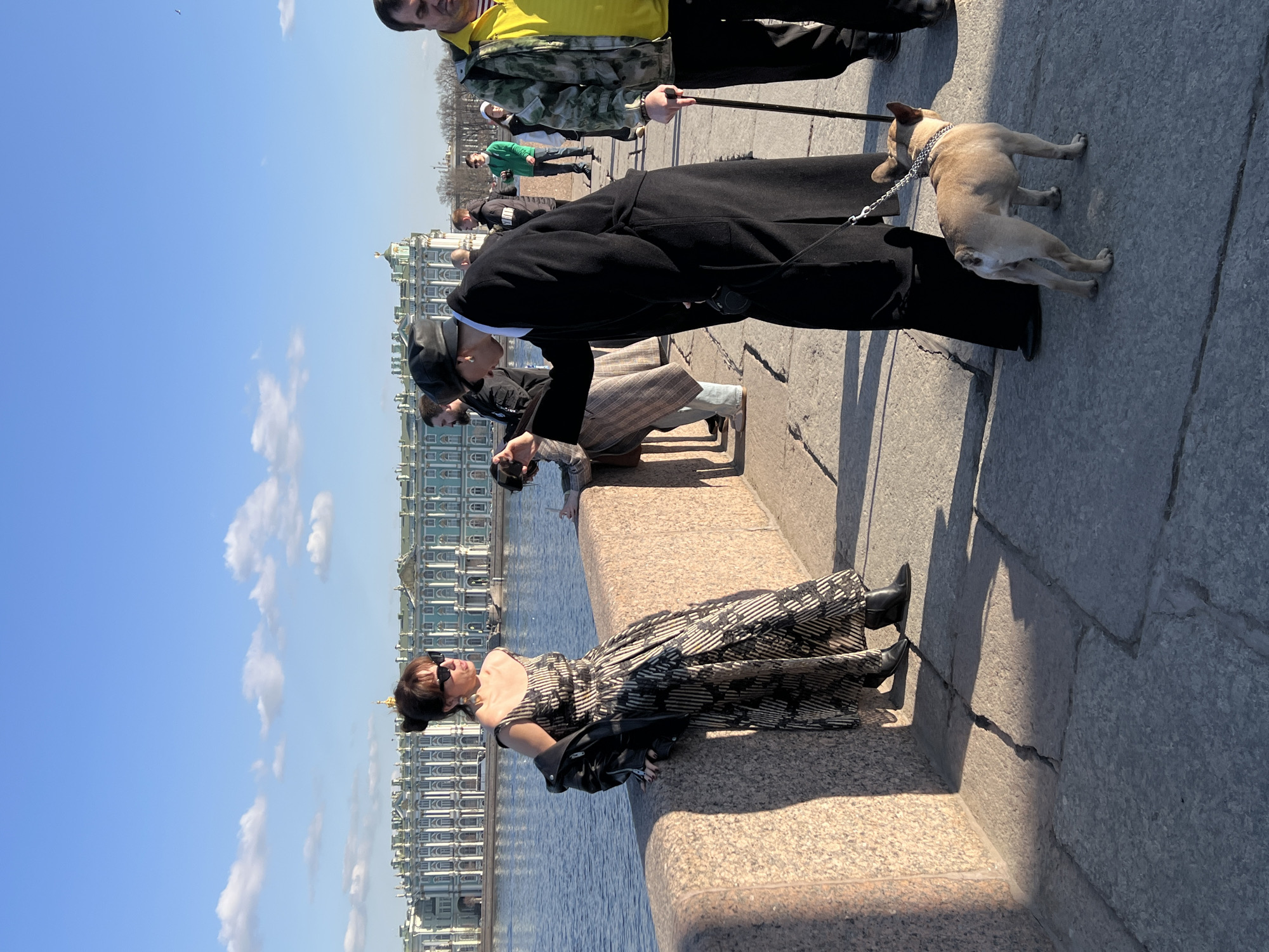 The image depicts an outdoor scene on a sunny day in Saint Petersburg, Russia, observed from the Palace Bridge. The Neva River flows to the left, with the turquoise and white Winter Palace (Hermitage Museum) prominently visible across the water in the background.
In the foreground, a woman with dark hair in a bun, wearing sunglasses, large earrings, a black and beige/gold patterned strapless dress, and a black leather jacket draped over her shoulders, leans against a granite-like stone parapet. She wears black boots and is looking to her right.
In the midground, a person dressed in a long black coat, a black beret, and sunglasses leans forward, looking down at a light tan French Bulldog on a leash. The person holds a small, dark object, possibly a camera or phone, in their right hand. The French Bulldog wears a chain collar and stands on the gray stone pavement.
To the right of this pair, a man in a camouflage-patterned jacket and a yellow t-shirt walks past, holding a dark stick or cane. Additional pedestrians are visible further in the background on the bridge. The sky is clear blue with scattered white clouds, and sunlight casts long shadows on the stone pavement.