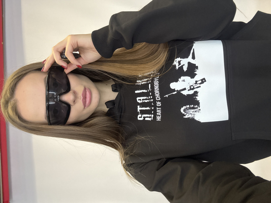 A young woman with long, straight, brown hair and red nail polish wears large black sunglasses and a black sweatshirt. The sweatshirt features a white graphic square with the words "STALKER HEART OF CHERNOBYL" and an image of people and objects in a wilderness. The woman is posed looking at the camera with a neutral expression. Her lips are light pink. A red pole is visible on the far left of the shot, and a light colored wall is in the background.