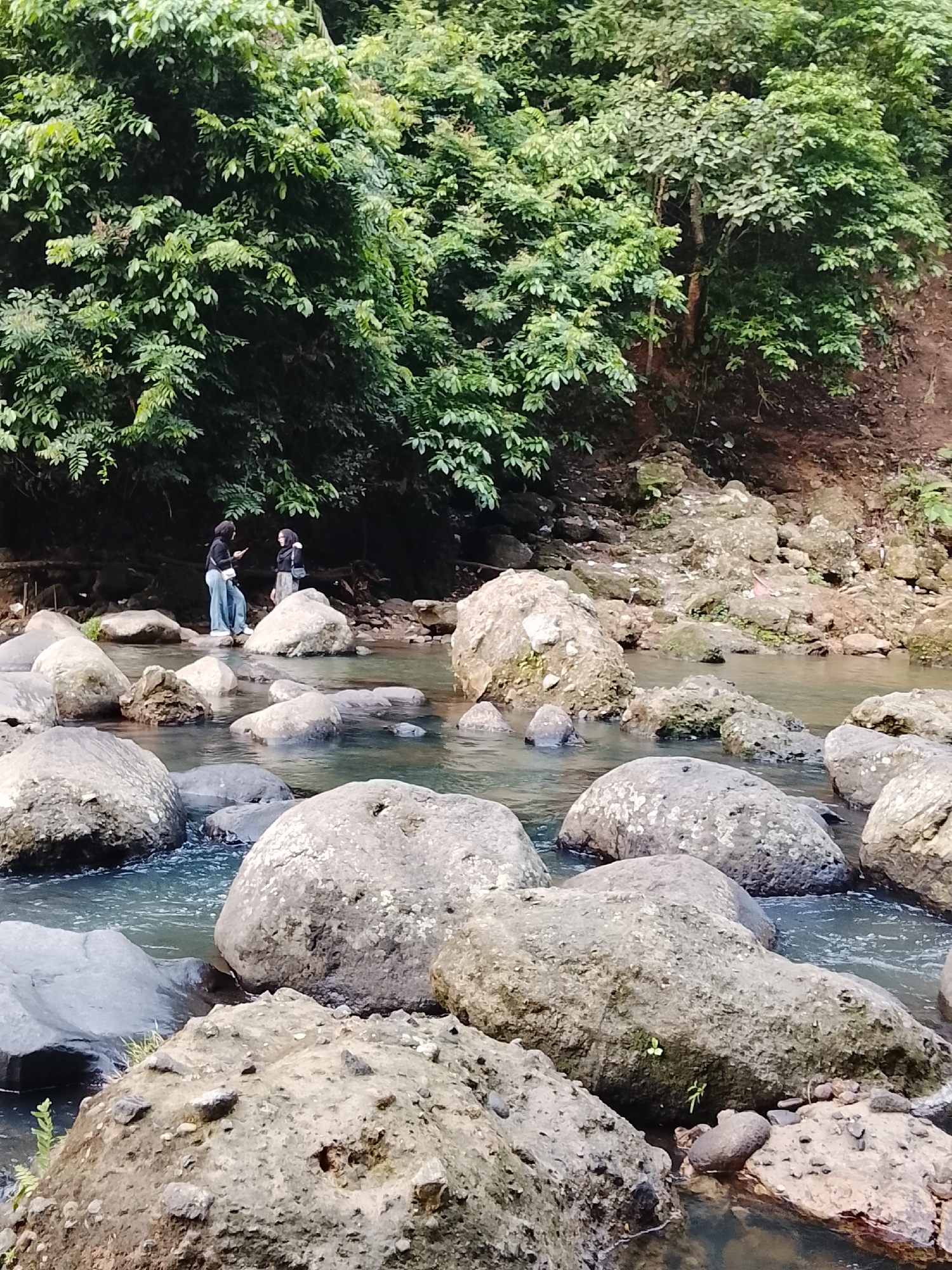 The image depicts a natural scene with a river and lush greenery. In the middle ground, two individuals are standing on rocks in a shallow river. One person is wearing a black hijab, black top, and light blue pants, while the other is wearing a black hijab and a skirt. They appear to be interacting with their mobile phones. The river is surrounded by large, rounded rocks of varying sizes, and the background is filled with dense green foliage and trees. The overall setting suggests a natural, outdoor environment, possibly a riverbank or a small waterfall area. The scene is consistent with the natural landscapes found in and around Lhokseumawe, Indonesia, known for its natural beauty and coastal areas.