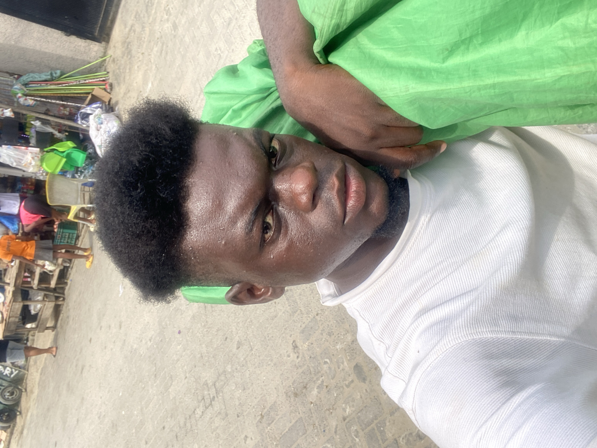 The photograph shows a man with dark skin, short black hair, and a white t-shirt. He appears sweaty, with beads of moisture visible on his face. He is holding what appears to be a green cloth over his shoulder. The background shows a street scene with stalls and people, suggesting an urban environment. The exact location is not specified.