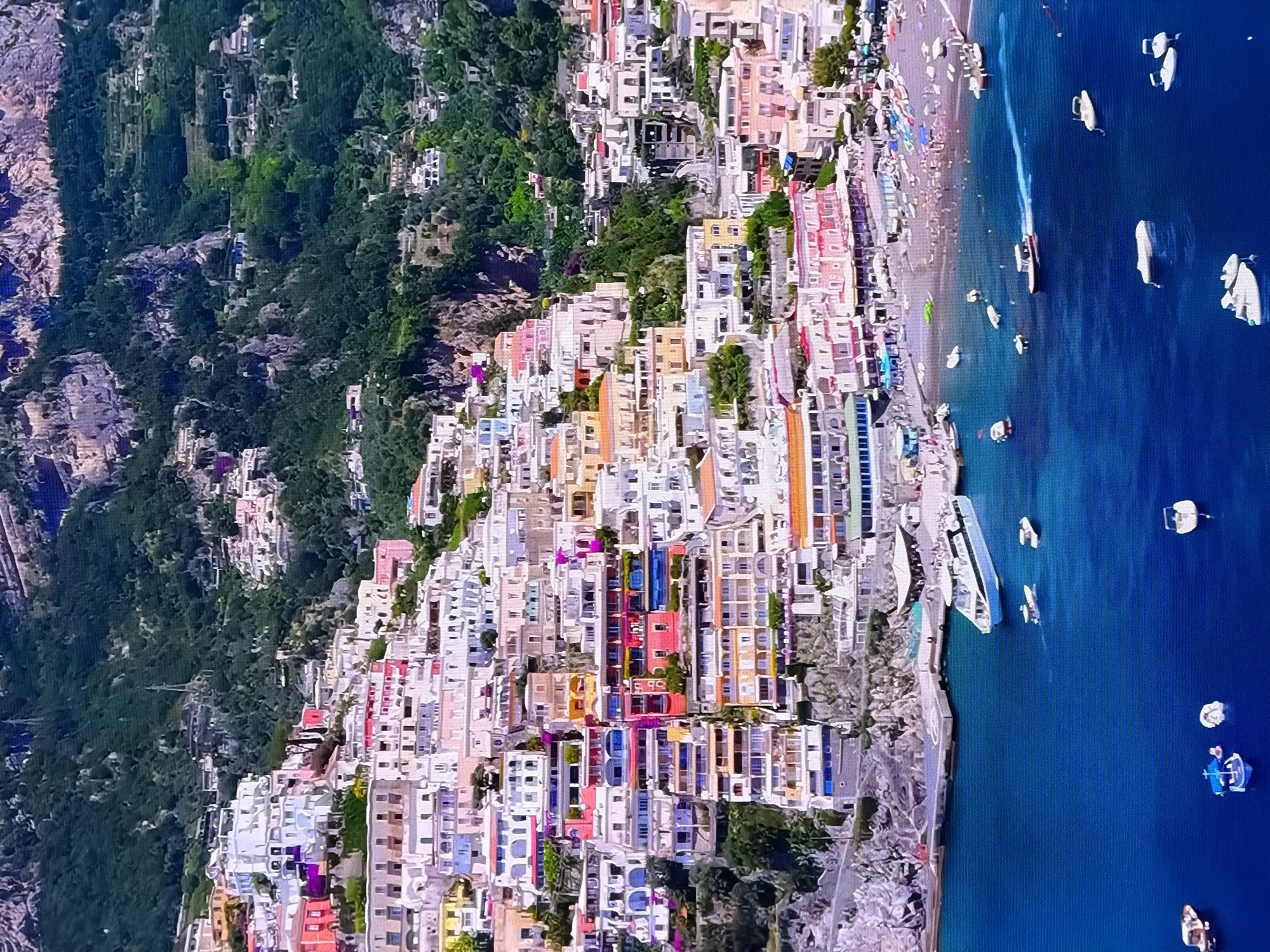 The image displays a coastal town nestled against a steep, verdant mountainside. Buildings of various colors, primarily white, pink, and pastel hues, are stacked tightly together, clinging to the slopes. Terraces and balconies are visible on many structures. A beach lined with umbrellas and loungers is situated at the base of the town, bordering a bright blue body of water. Several boats, including a large ferry and smaller vessels, are docked or moving in the water. The sky is not visible in the frame, and the overall impression is that of a sunny day in a picturesque Mediterranean or similar coastal location. The request to include the city of Chirchik, Uzbekistan, is not supported by the visual content of the image.