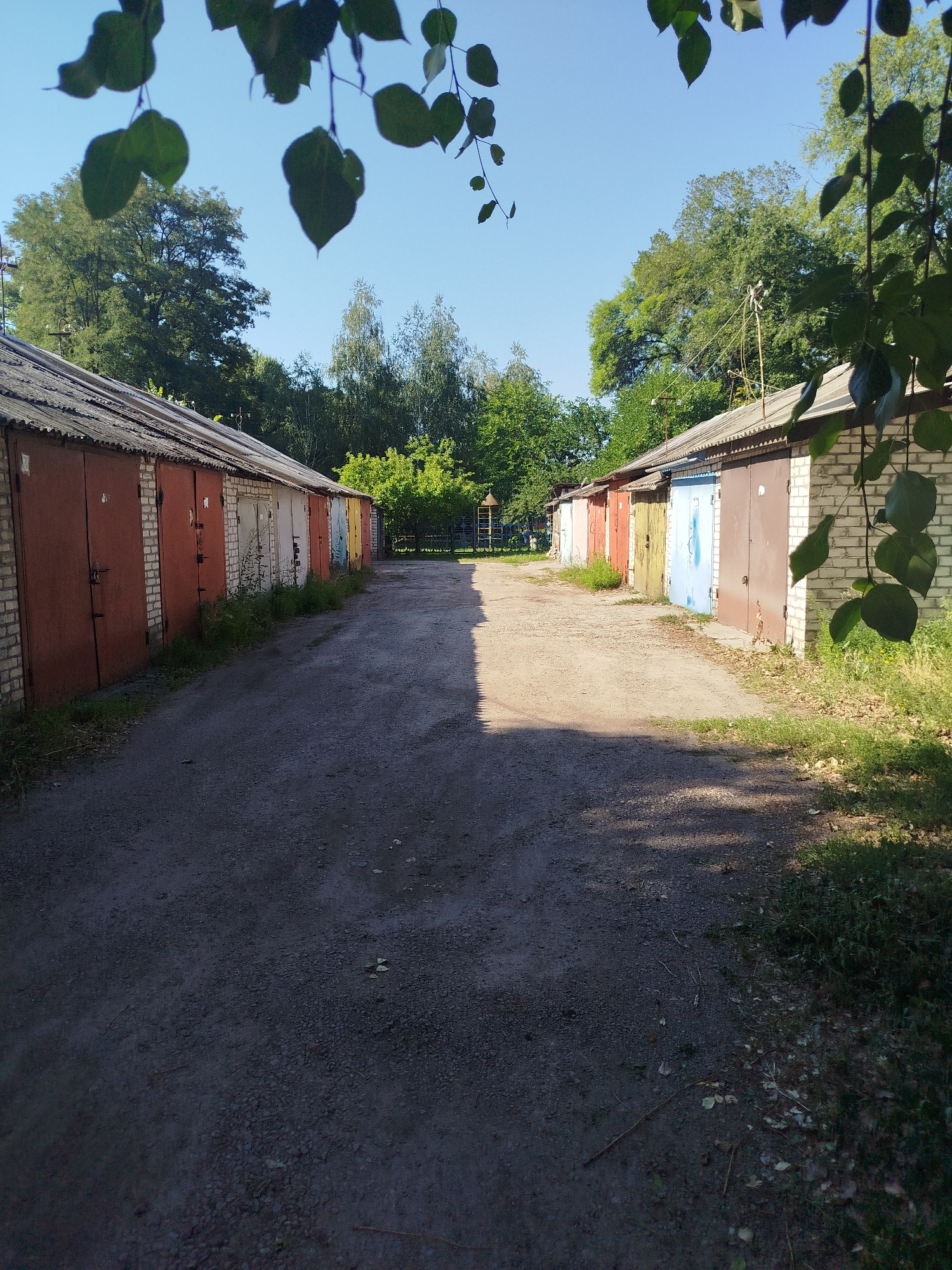 The image shows a dirt road between two rows of garages in Kremenchuk, Ukraine. The garages are made of brick and metal, with corrugated metal roofs. The doors of the garages are various colors, including red, blue, and brown. Trees and green foliage are visible in the background. The sky is blue and clear, indicating a sunny day. The road is unpaved and appears to be well-traveled, with tire marks and scattered debris. The overall impression is one of a utilitarian and somewhat neglected area, typical of a garage complex in a residential or industrial district.