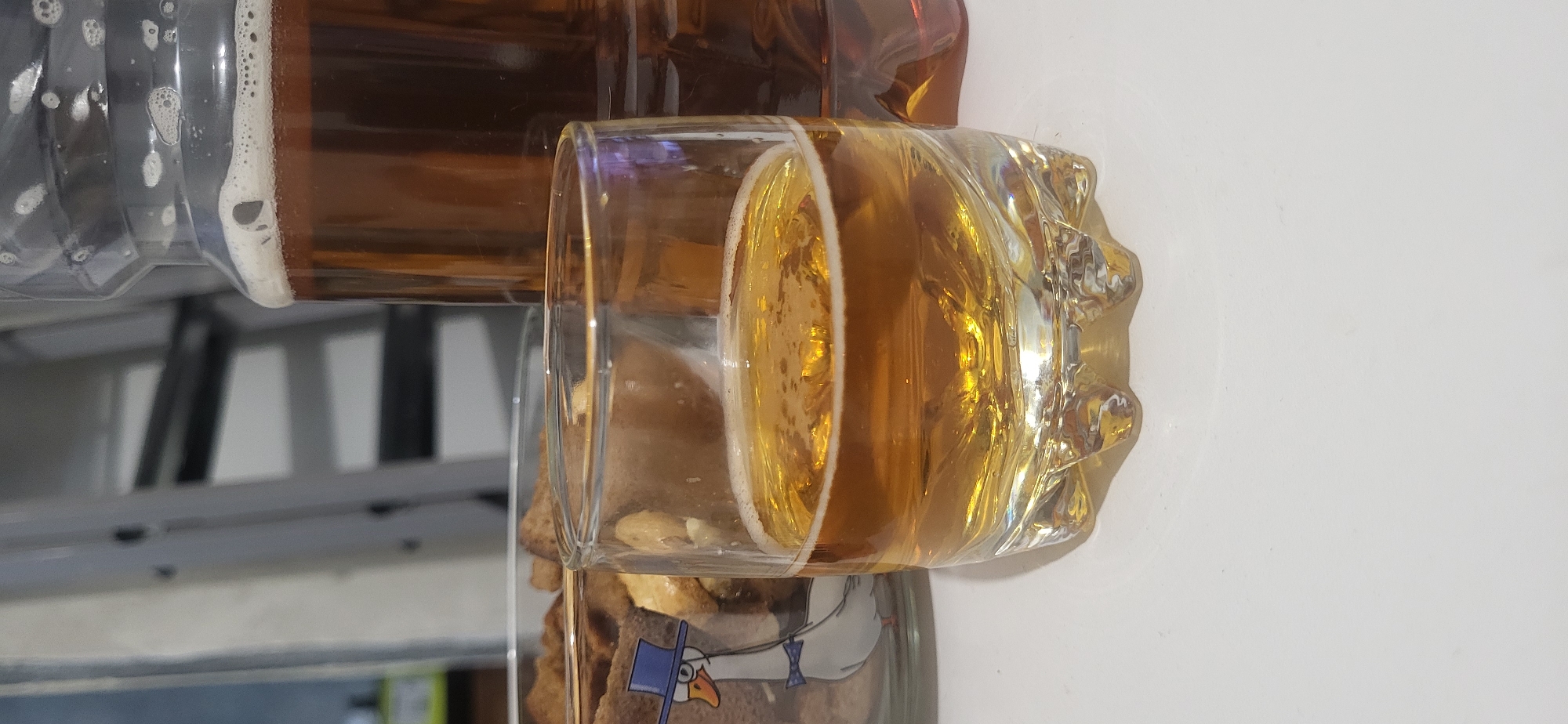 The image shows a close-up of a glass of beer being poured from a bottle. The beer is a light amber color with a white foam head. The glass has a textured exterior. The bottle appears to be clear plastic. The scene takes place in the city of Unknown, Unknown. In the background, what appears to be a shelf and a jar containing food are visible.