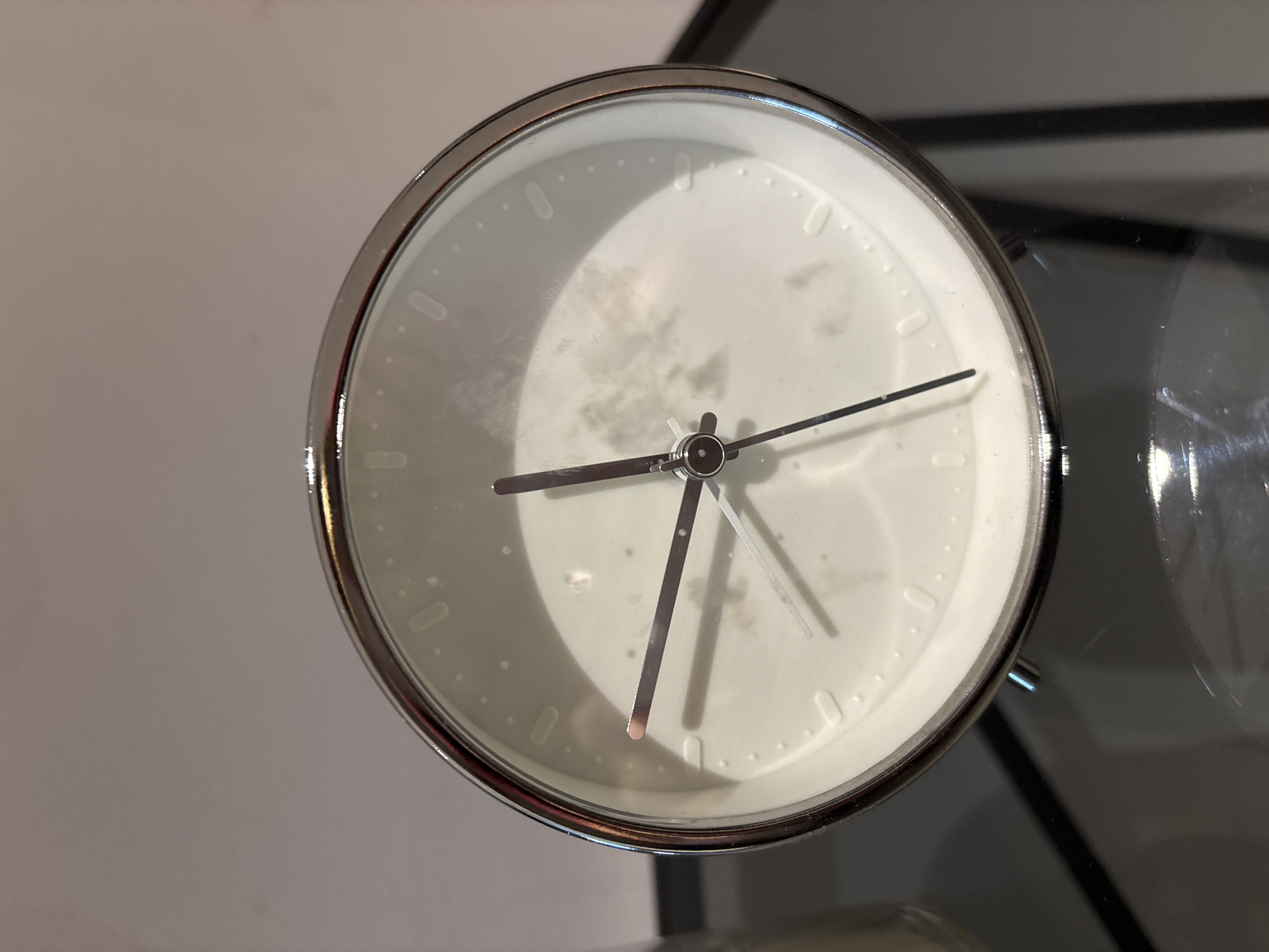 A close-up, eye-level, medium shot shows a white analog alarm clock with a silver frame resting on a dark, reflective surface. The clock face has simple hour markers represented by small dots and longer dashes, with the numbers 12, 3, 6, and 9 being more prominent. The hour hand points to approximately 10, and the minute hand points to approximately 2. A thin second hand is also visible. The clock face has some smudges and imperfections. The background is a blurry, neutral color. The scene is indoors and appears to be a still life composition. No specific location in Dubai, United Arab Emirates, can be determined from the image.