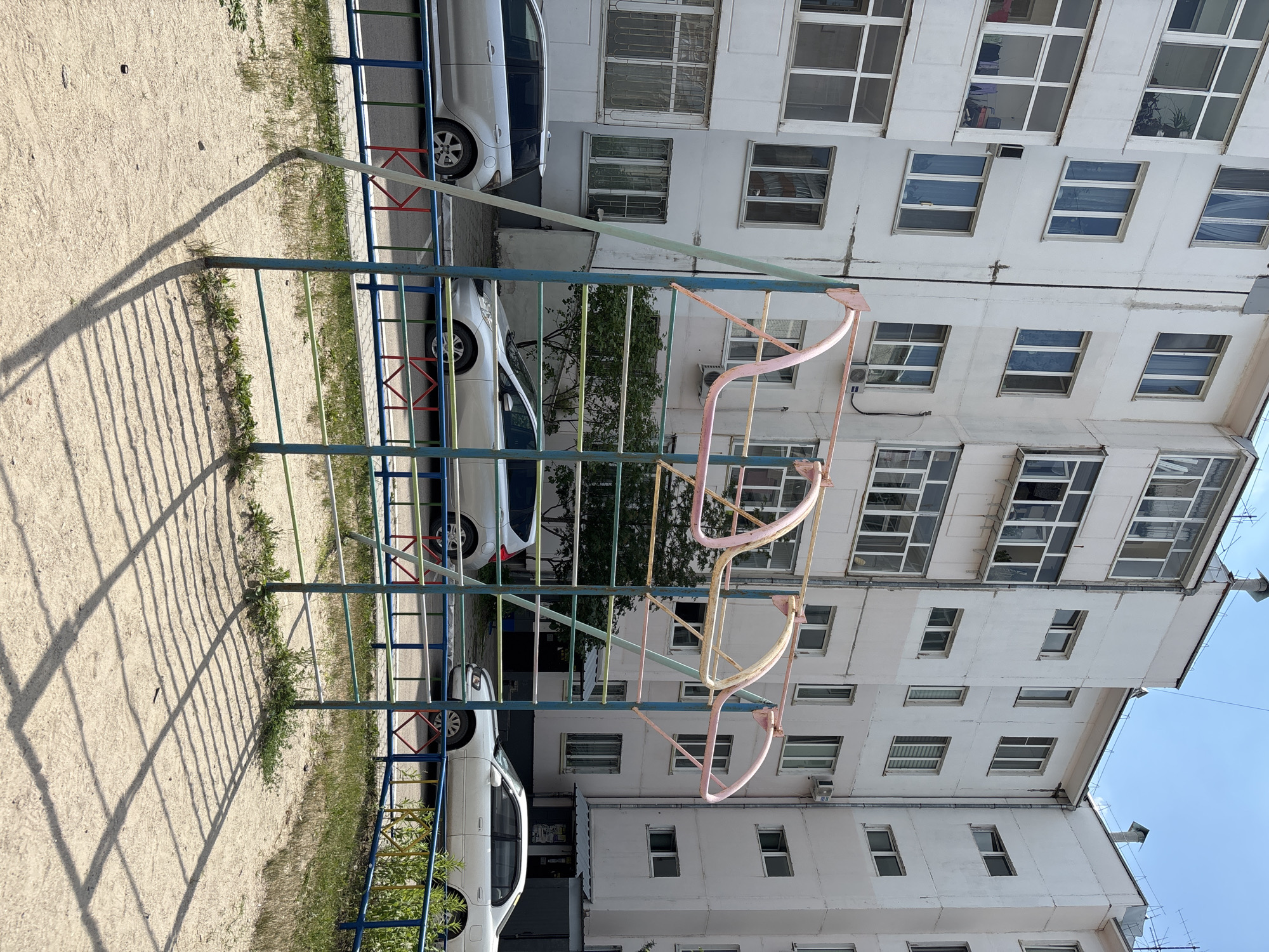 In Blagoveshchensk, Russia, a bright, sunny day reveals a residential courtyard with a playground and parked cars. A white, multi-story apartment building with many windows forms the backdrop. In the foreground, a colorful metal playground structure with pink and yellow bars stands on a sandy ground. Two silver cars are parked on the asphalt next to the playground, separated by a blue, red, and green metal fence. Green grass and plants grow along the fence line.