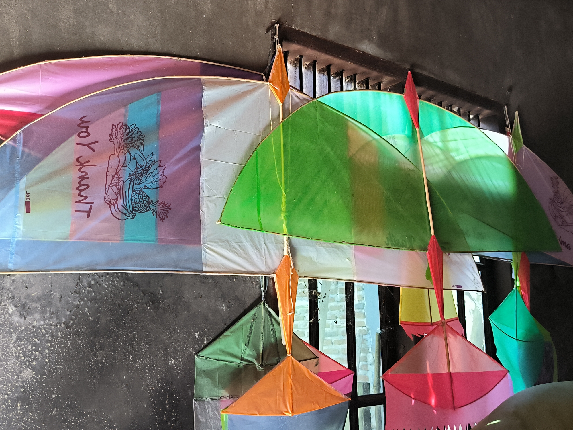 The image shows a collection of colorful kites hanging from a dark-colored ceiling fixture against a dark wall. The kites are of various shapes and colors, including green, orange, pink, and white. One kite has the words "Thank you" printed on it, along with an image of vegetables. The background reveals a window with a view of a brick building. The location is Merbopuntong, Indonesia.