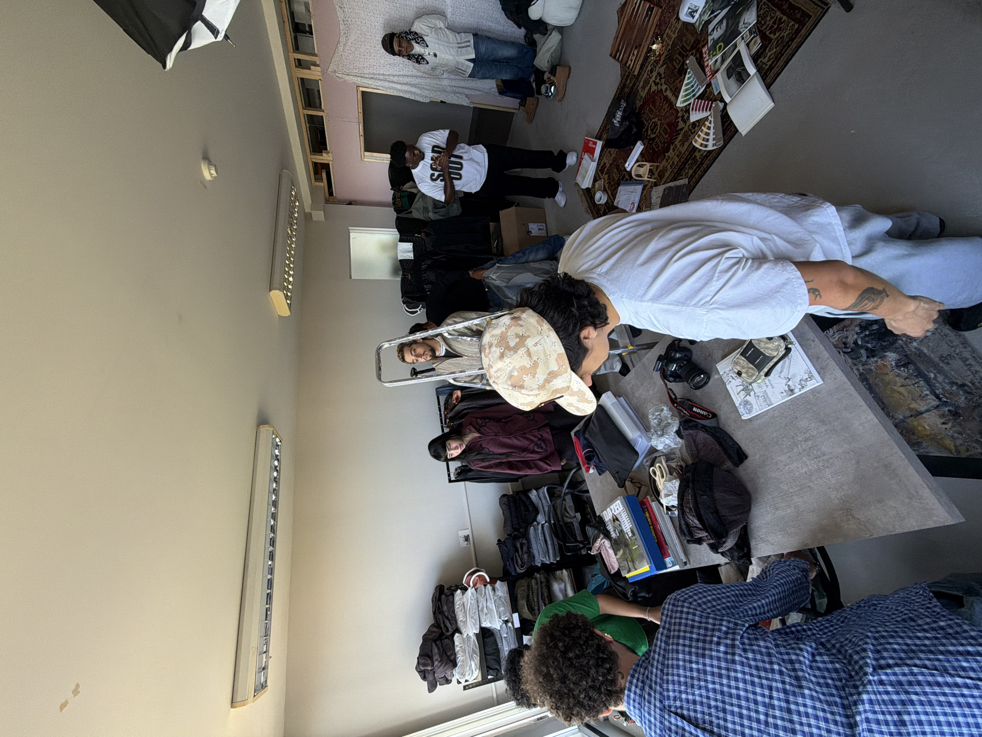 This image, rotated 90 degrees clockwise, captures a dynamic indoor scene, likely a creative studio or retail space in London, United Kingdom. Approximately seven to eight individuals are present, engaged in various activities.
In the foreground, the back of a person wearing a blue and white checkered shirt is visible, alongside another in a green t-shirt. To their right, a person with a camouflage cap and a white t-shirt, adorned with arm tattoos, leans over a large grey table. This table holds a black Canon camera, lenses, papers, scissors, books, and other items, suggesting a work or photography setup.
Further into the scene, a person in a white t-shirt clearly displaying "SOLD OUT" in black text stands prominently. Nearby, another individual in a burgundy top and black headwear is visible. Towards the back, a person in a white jacket and blue jeans stands beside what appears to be a low bed or platform, while another leans on a metal structure or step-ladder.
The room features light grey walls and a dark grey floor, partially covered by a patterned rug. Multiple clothes racks brimming with various garments (jackets, hoodies, bags) are arranged throughout the space, reinforcing the idea of a fashion-related environment. Overhead, a black softbox, typically used for photography lighting, is visible in the upper left corner. Fluorescent light fixtures are mounted on the walls. A small white object, possibly a smoke detector, is on the ceiling. The overall atmosphere appears industrious and casual, with people gathered, possibly collaborating or preparing for an event.
