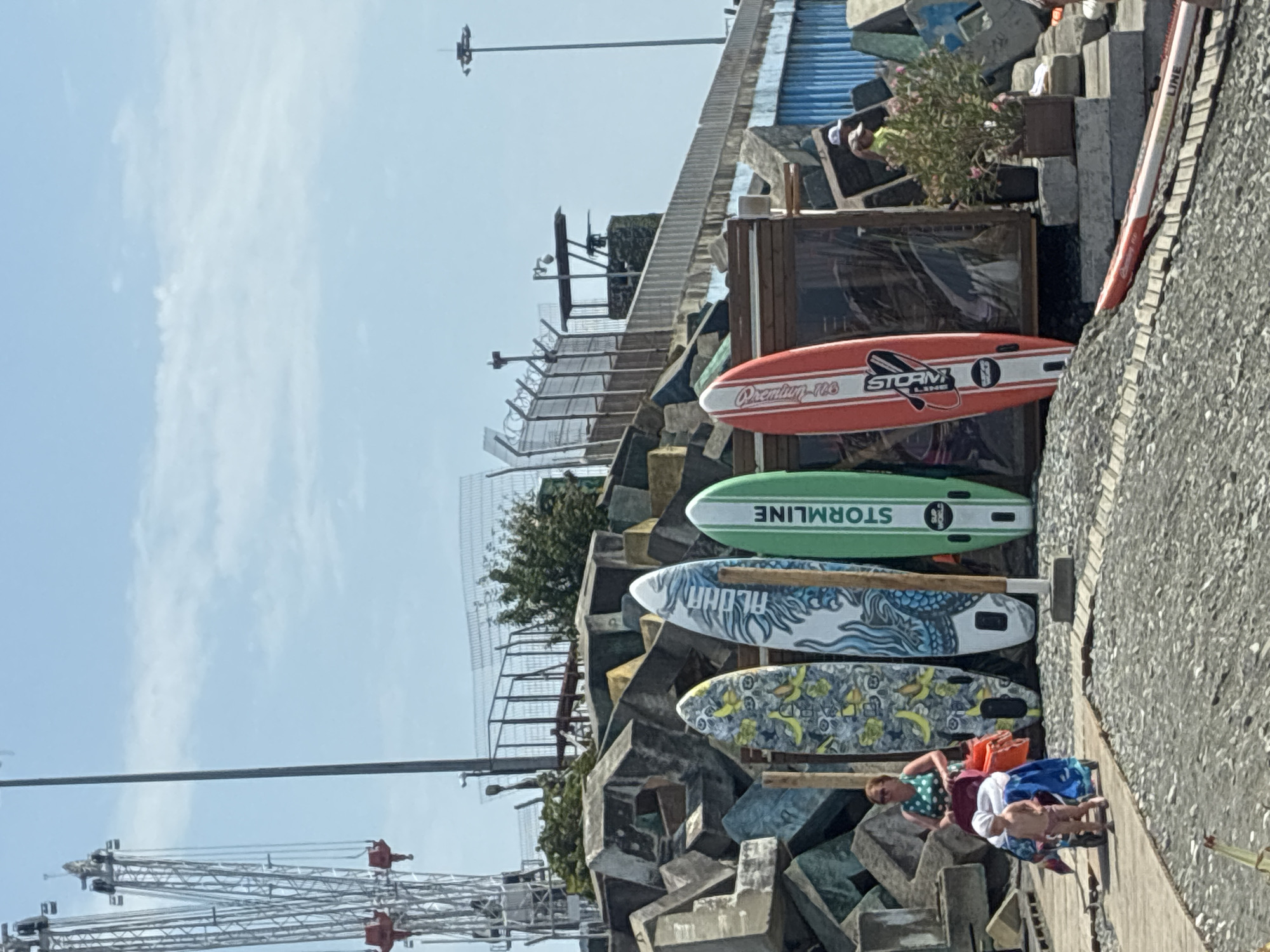 The image depicts an outdoor scene with a clear blue sky and a few wispy clouds. In the foreground, there is a wooden boardwalk leading to a pebbled area. Several surfboards and stand-up paddleboards are propped up against a structure made of large, angular concrete blocks. One surfboard has a banana and yellow butterfly pattern. Another is white with a blue dragon design. A green board has the text "STORMLINE" and "SUP WORLD" printed vertically. An orange and white board also features the "STORMLINE" logo.
To the left of the surfboards, a woman in a green polka-dot top is pushing a stroller containing a baby. The baby is wearing a white bonnet and appears to be in a pink outfit. Behind the woman and the boards, a complex industrial structure with cranes and scaffolding is visible against the sky. Further back, a fenced area with guard posts and security cameras suggests a restricted zone. In the background, a tall light pole stands to the right.
To the right of the surfboards, there is a small wooden kiosk or shop with a transparent screen. A person wearing a yellow shirt and a white cap is standing near it, along with a potted plant with pink flowers. Another individual, possibly wearing a light-colored hat, is partially visible. A second stand-up paddleboard is lying on the ground in the lower right corner of the image. The overall impression is a recreational area adjacent to industrial infrastructure, possibly near a port or coastal facility. The context of Sirius, Russia, suggests this may be a location within that coastal resort city.