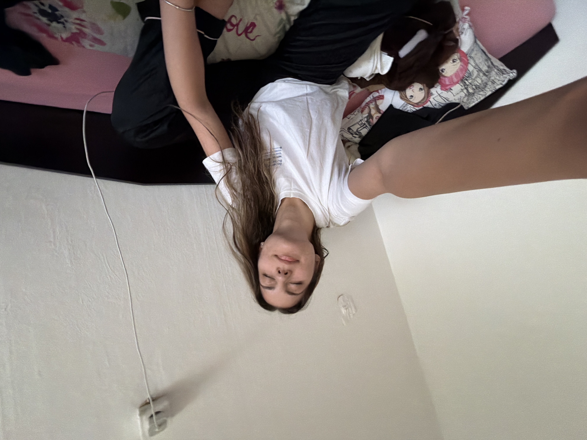 A woman is lying on a bed in a room. She has long brown hair and is wearing a white t-shirt and black pants. The bed has pillows with designs on them. There is a white cord running along the wall. No information is provided to place this location in Sofia, Bulgaria.