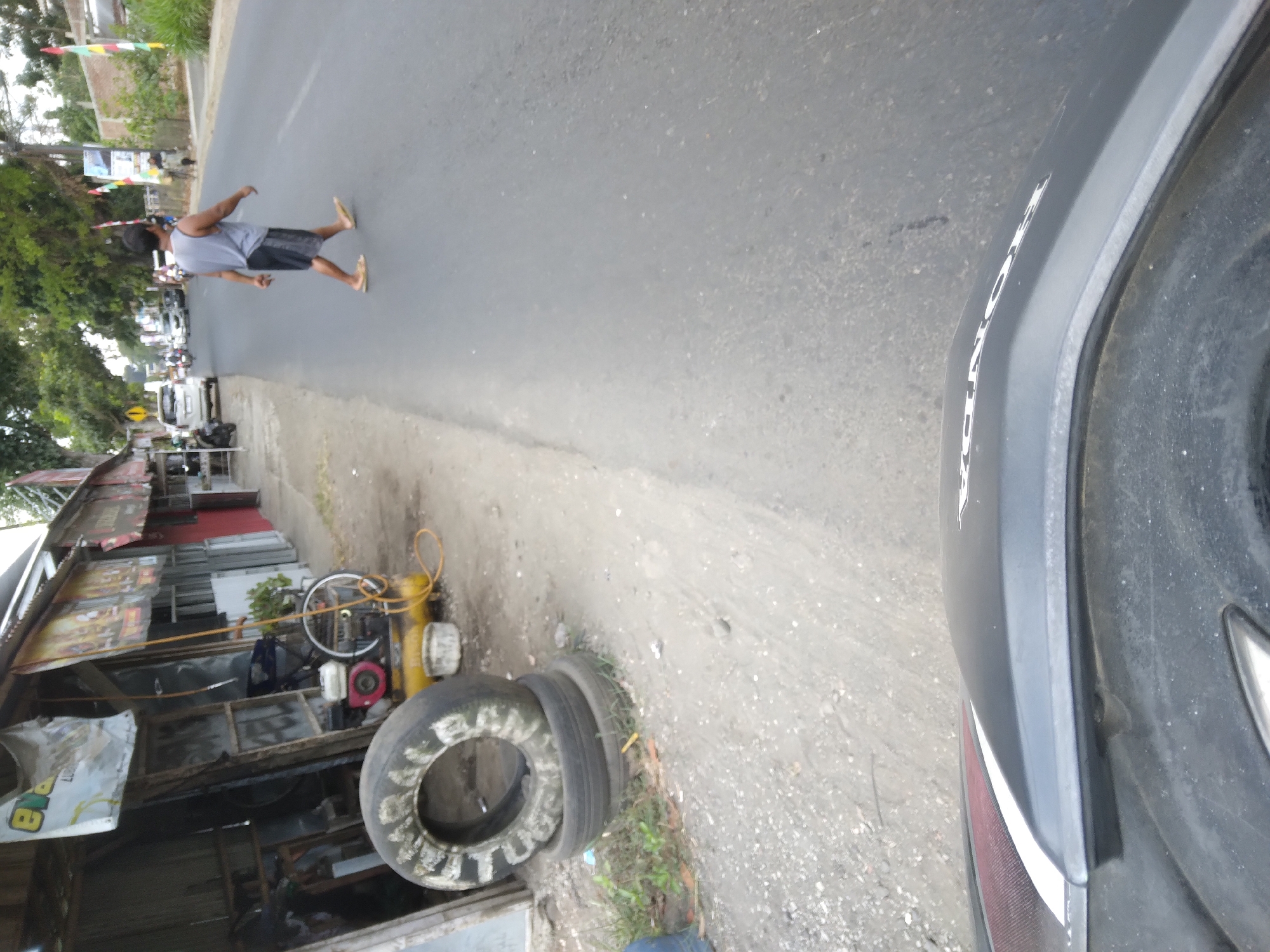 This is a street-level photograph taken in Sokaraja, Indonesia, likely from the perspective of a moving vehicle. The scene captures a stretch of asphalt road bordered by a dirt shoulder and several roadside structures. In the foreground, the curved black plastic body of a motorcycle is visible, suggesting the vantage point of a rider.
On the left side of the road, a small shop or workshop is partially visible, featuring a corrugated metal roof and open-air shelving. Stacked tires, one significantly larger than the others, are positioned on the dirt shoulder in front of this establishment. Adjacent to the tires, a yellow air compressor with a red engine and a blue tank is present, along with a yellow hose. Further back, a bicycle is leaning against a support beam.
The road itself is a two-lane asphalt surface. A lone individual, a male wearing a grey sleeveless tank top and dark shorts, is seen walking across the road from right to left. He is barefoot. Several vehicles, including cars and motorcycles, are traveling on the road in the background, indicating moderate traffic.
The environment suggests a warm, possibly humid climate, with lush green trees lining the road in the distance. The roadside also features various flags and banners, suggesting a populated or commercial area. The overall impression is of a typical Indonesian roadside scene, likely in a town or semi-urban area.