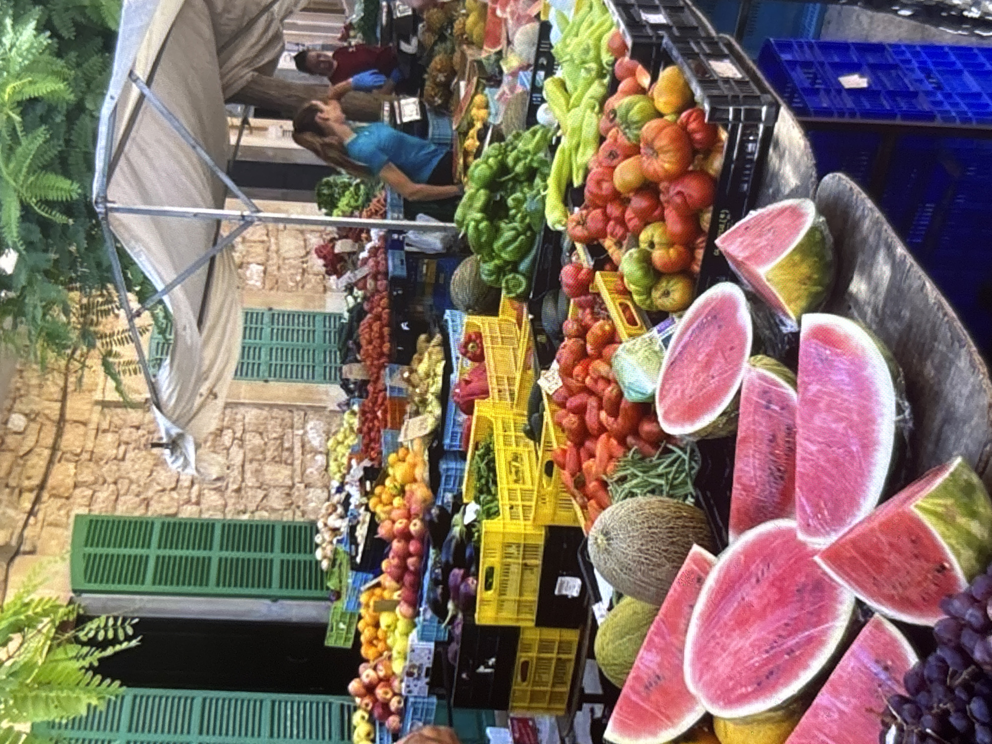 The image depicts an outdoor market scene, likely in Orihuela, Spain, characterized by stalls overflowing with fresh produce. In the foreground, sliced watermelons and a bunch of grapes are prominently displayed on wooden surfaces. Behind these, various fruits and vegetables are arranged in crates and baskets. Among the produce, one can identify tomatoes, melons, green peppers, and a variety of other fruits like apples and oranges. Two women are visible in the background; one in a blue top is attending to the produce, and another in a red top appears to be assisting. The market is set against a backdrop of a stone building with green shutters, suggesting an old-world architectural style typical of Spanish towns. A white canopy provides shade over the market stalls. The overall impression is one of a vibrant and bountiful local market.