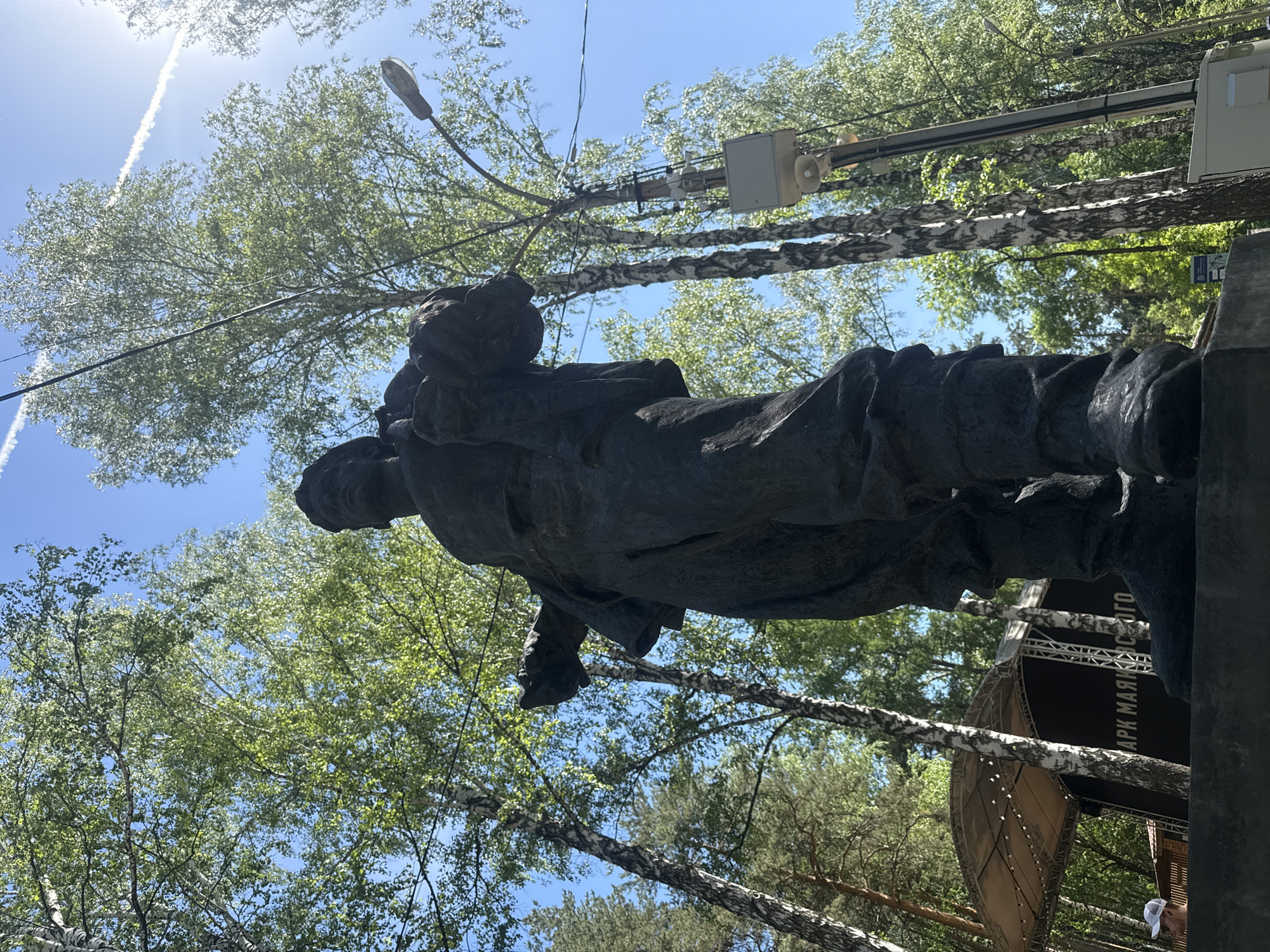 The image shows a low-angle view of a dark statue against a backdrop of trees and a blue sky with contrails. The statue is a man in what appears to be working clothes, possibly holding something. In the background are birch trees, utility poles, and a light fixture. A stage-like structure is partially visible behind the statue, with the text "АРК МАЯКО С ОГО". The location appears to be the Mayakovsky Park in Yekaterinburg, Russia.