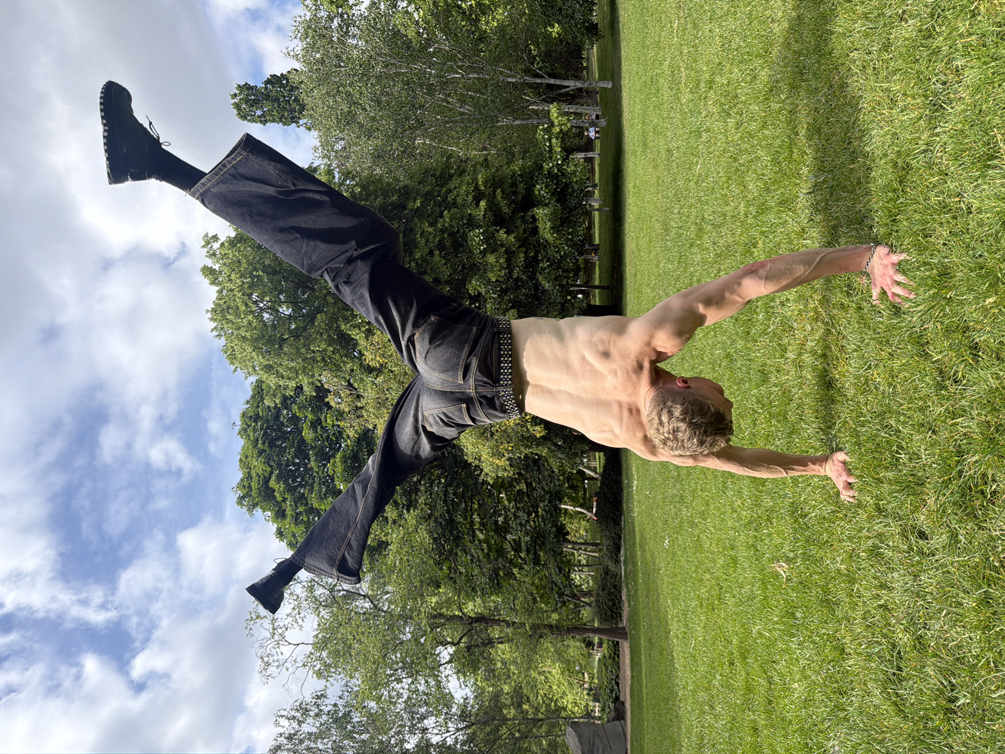 A shirtless man wearing dark jeans with black boots and a studded belt is performing a handstand in a park. The grass is bright green and there are trees in the background. The sky is visible above the trees, with blue patches and some clouds. The photograph is rotated 90 degrees counterclockwise.