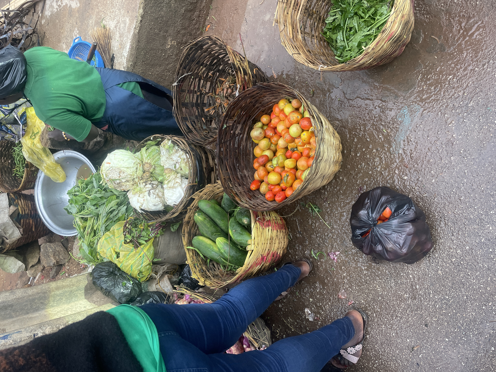 This image depicts a street market scene in Jos, Nigeria. The ground is wet, suggesting recent rain. In the foreground, a person wearing blue jeans and a green top stands near various produce. To the left, there are bags of vegetables. A large wicker basket filled with cucumbers is prominent. Adjacent to it, another basket overflows with red and yellow tomatoes. Further right, a basket contains green leafy vegetables. Behind these, larger baskets hold cabbages and what appear to be root vegetables. A person wearing a green t-shirt and a dark blue apron is visible in the background, interacting with the produce. The overall environment suggests a bustling local market.