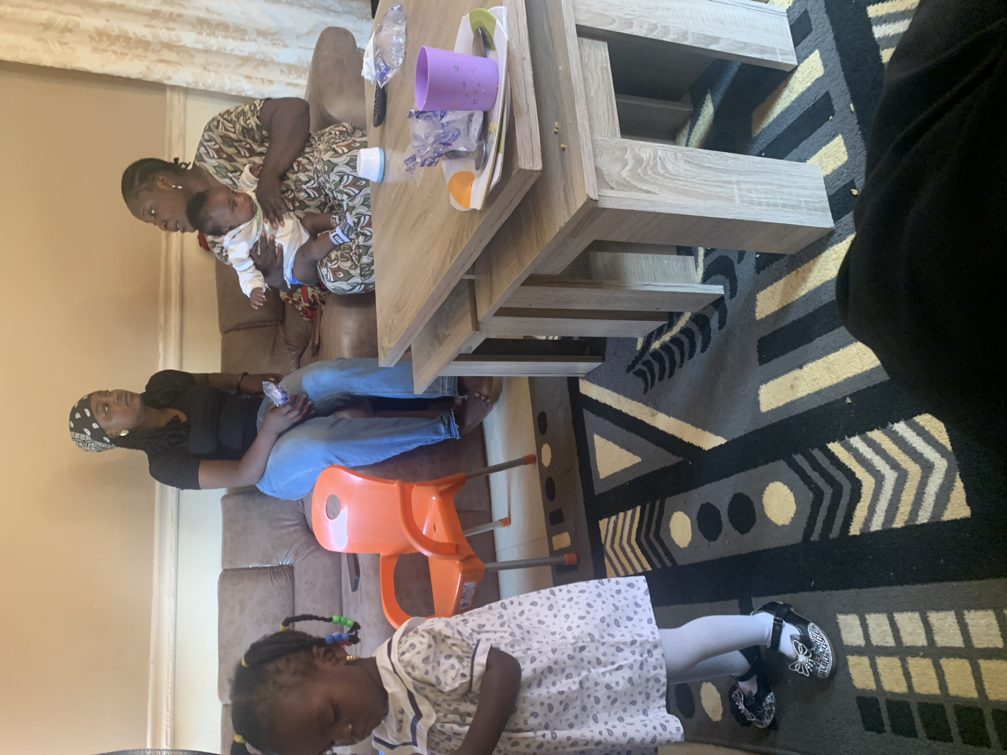 In a domestic interior, possibly in Gura Top, Nigeria, two women and two children are present. The scene appears to be a living room.
On the left, a young girl stands facing away from the camera, wearing a light-colored dress with a patterned print and white tights, accessorized with black shoes adorned with bows and rhinestones. Her hair is styled in two braids, decorated with colorful beads.
To her right, an orange plastic child's chair is visible. Further back, on a brown sofa, a woman wearing a black t-shirt and blue jeans sits, holding a clear plastic bottle. To her right, another woman is seated on the sofa, holding a baby. The baby is dressed in a white onesie and blue bottoms. The woman holding the baby wears a patterned dress and a headscarf.
A light-colored wooden coffee table is in the foreground, positioned on a rug with a geometric design in black, grey, yellow, and white. On the table, there is a plate with food remnants, a purple cup, a remote control, and a packet of tissues.
The environment suggests a casual, indoor setting with furniture including a sofa, a coffee table, and a child's chair. The overall lighting is moderate.