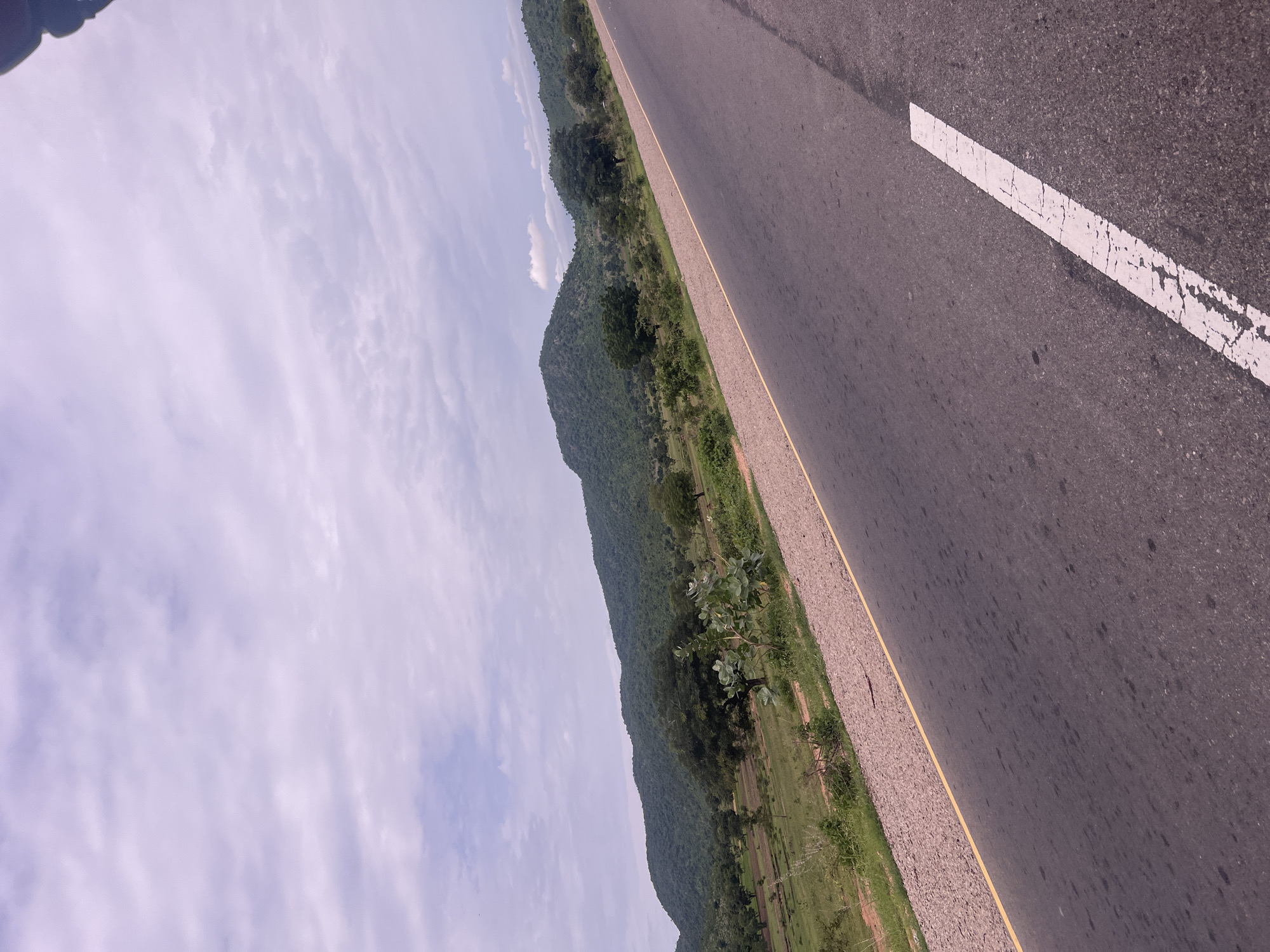 This media file depicts a roadside view in Minda, Nigeria. The scene is dominated by a dark asphalt road running diagonally from the lower right to the upper left corner of the frame. A broken white line, characteristic of road markings, is visible on the right side of the road. A yellow line also demarcates the edge of the road. To the left of the road, a gravel shoulder transitions into a lush green landscape. Rolling hills covered in dense vegetation rise from the green fields. The sky above is filled with light, wispy clouds, suggesting a bright day, possibly late morning or early afternoon. The overall impression is one of a rural or semi-rural setting, highlighting the road as a passage through a natural, verdant environment. There is no visible text in the image. The perspective suggests the image was taken from a moving vehicle.