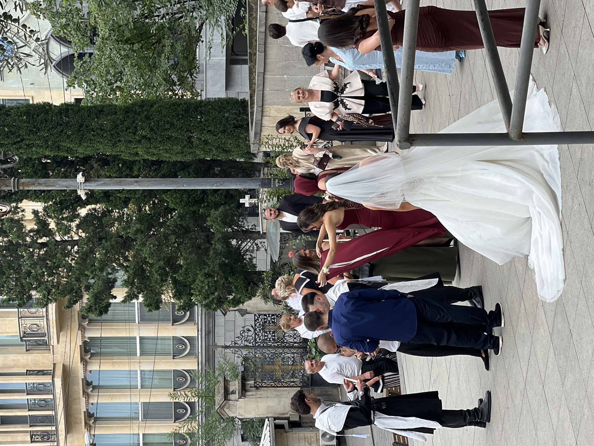 The image captures a wedding party gathered outside a grand building in Tbilisi, Georgia. The architecture suggests a historical district, likely near an orthodox church, given the presence of a cross visible on a fence in the background and the ornate iron gate. Several people, dressed in formal attire, are interacting. A bride in a white gown and veil stands prominently on the right, with guests surrounding her. Some men are dressed in what appears to be ceremonial robes, possibly part of a wedding procession or tradition. The environment is an outdoor urban setting with paved ground and trees providing a natural backdrop to the stone facade of the building.