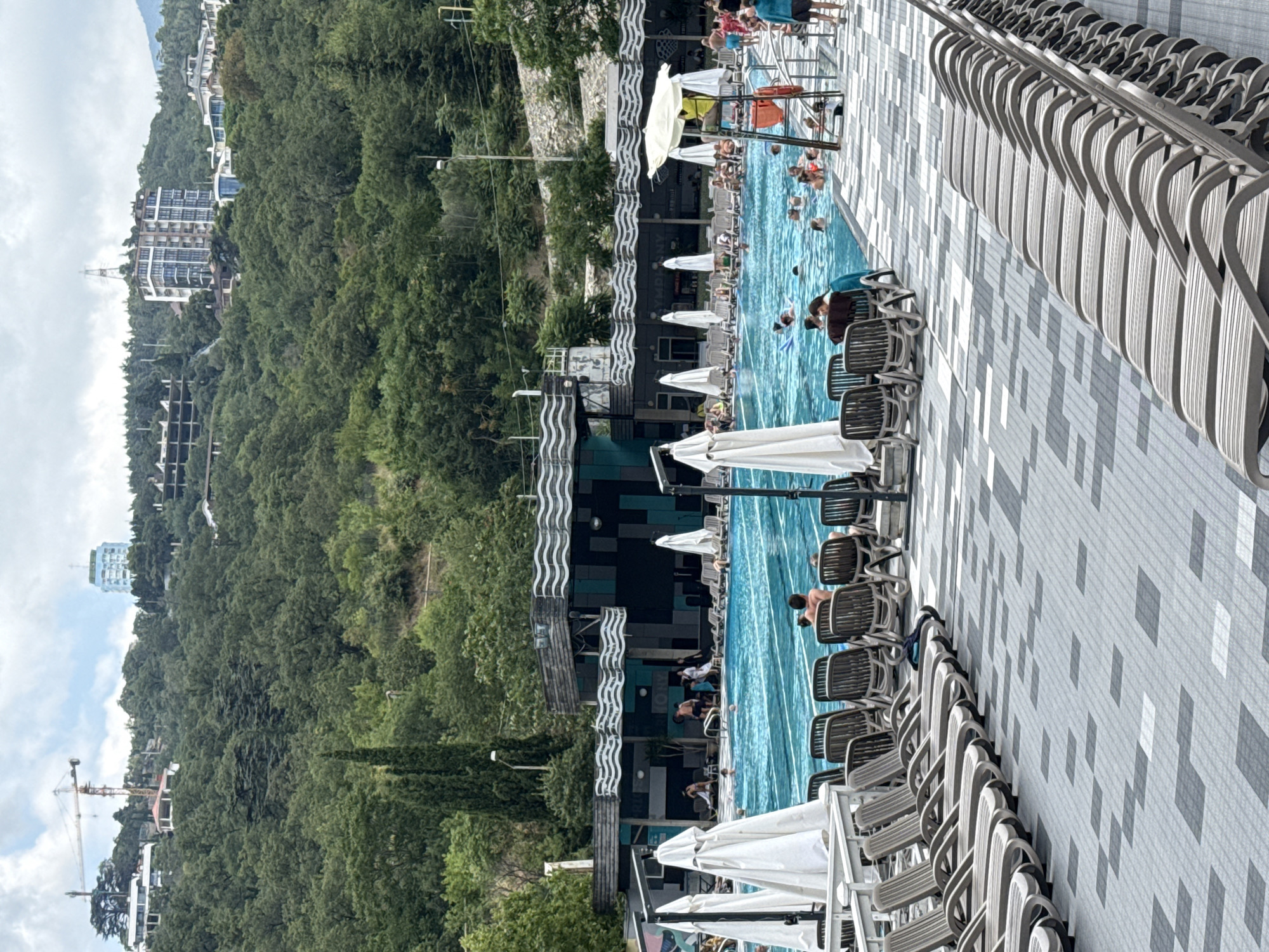 The image depicts an outdoor swimming pool area situated on a hillside in Yalta, Ukraine. The pool is a large, rectangular body of blue water with lane markings, and it is populated by numerous individuals swimming and relaxing. Surrounding the pool are rows of lounge chairs, many of which are occupied by sunbathers. Several large white umbrellas provide shade. In the background, a multi-story building with a dark facade and decorative wavy elements runs along the edge of the pool. Beyond this structure, a lush green hillside covered in trees rises steeply. Several buildings and a construction crane are visible on the upper slopes of the hill, indicating ongoing development or construction in the area. The sky is overcast with clouds. The overall scene suggests a recreational facility in a resort-like setting.