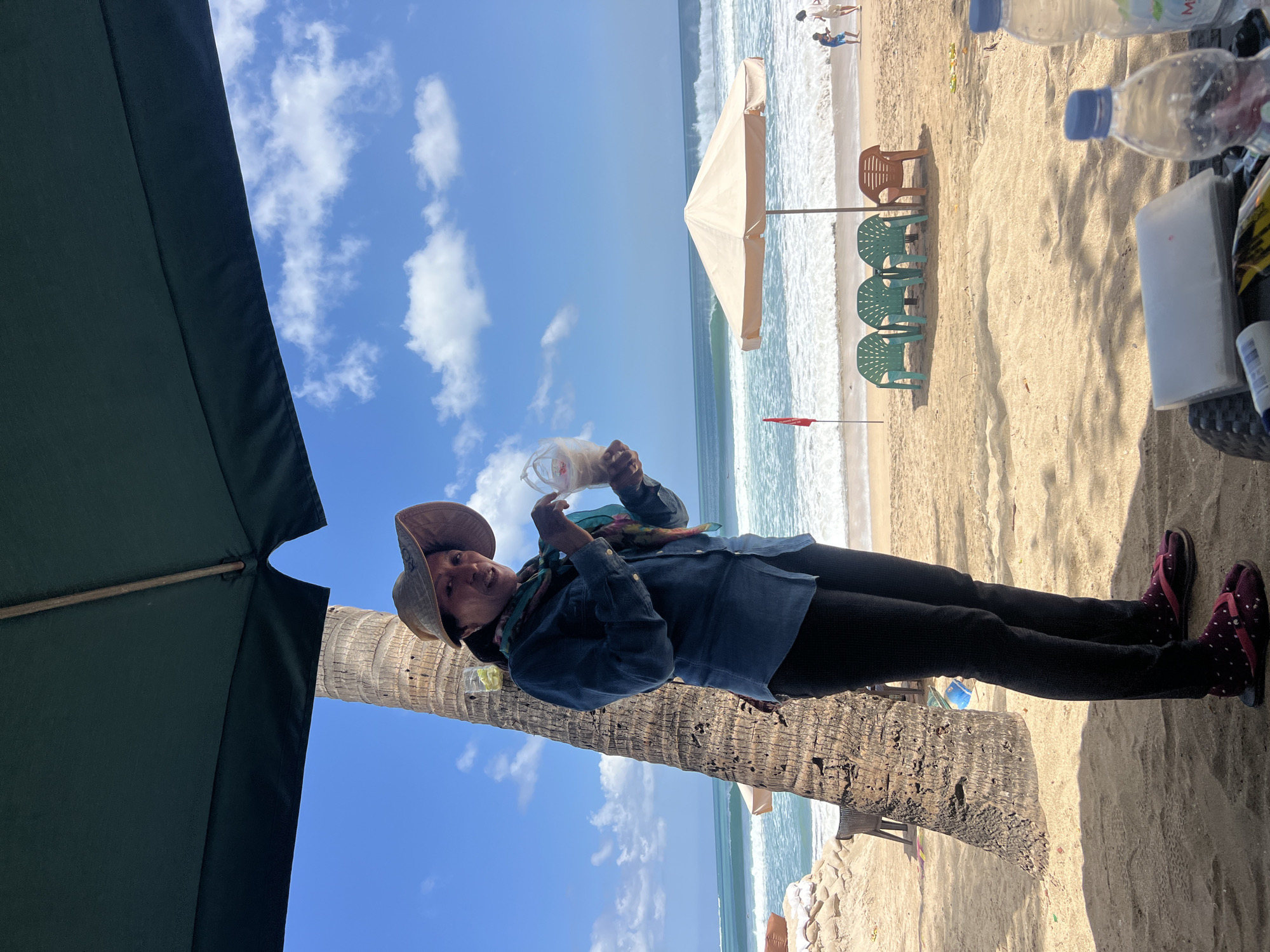 A woman wearing a straw hat, a blue button-down shirt, black leggings, and split-toe sandals stands on a sandy beach in Kuta, Indonesia. She is holding a clear plastic bag containing a white substance, possibly food or drink. Behind her, a palm tree stands tall, and the ocean waves crash onto the shore under a bright blue sky with scattered clouds. Beach chairs and an umbrella are visible further down the beach, where a few people are walking. The scene is illuminated by natural sunlight, casting shadows on the sand.