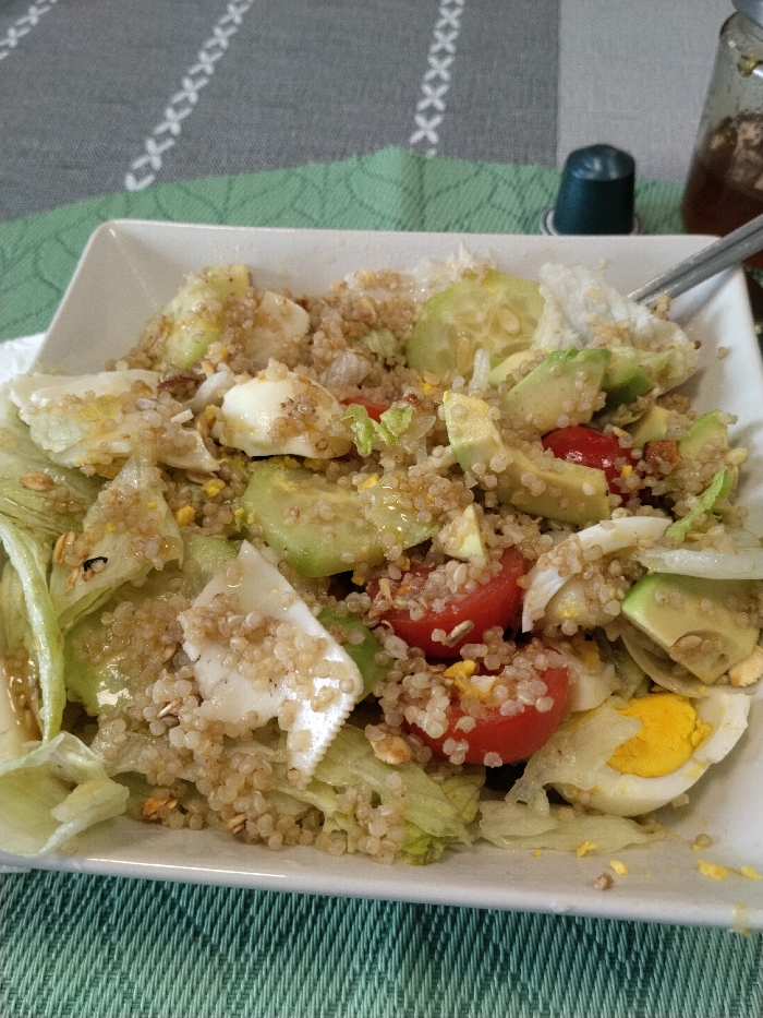 A white, rectangular dish contains a salad composed of quinoa, shredded green lettuce, halved red cherry tomatoes, sliced pale green avocado, sliced hard-boiled eggs revealing white albumen and yellow yolk, and sliced light green cucumbers. A clear, oily dressing coats the salad ingredients. A metallic utensil, consistent with a fork, is partially inserted into the salad on the upper right side. The dish rests on a textile tablecloth featuring a dual-tone design: dark grey on the left and light green on the right, separated by a vertical seam. Both sections of the tablecloth exhibit a repeating white chain-link pattern formed by 'X' shapes. In the background, behind the dish and to the right of the tablecloth's color divide, a dark green cylindrical object is present. Further to the right, a clear glass bottle containing a light brown liquid, with a metallic utensil resting inside, is partially visible. The image provides no discernible visual information to establish its location within the city of Vila Real, Portugal.
