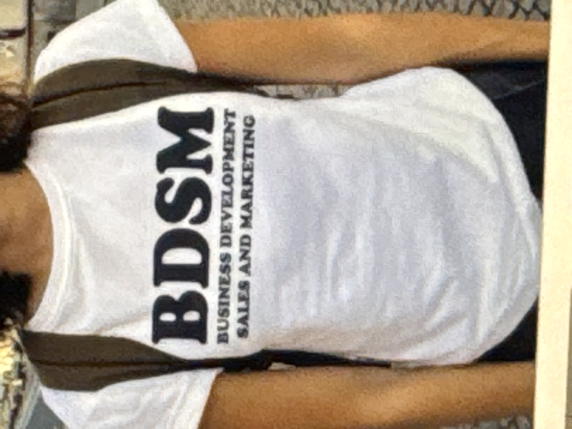 A person is wearing a white t-shirt with black text that reads "BDSM" in large font at the top, followed by "BUSINESS DEVELOPMENT SALES AND MARKETING" in smaller font. The person is also wearing a dark backpack. The image is a close-up shot of the t-shirt and the upper body of the person. The background is blurred and appears to be an outdoor urban setting, possibly a street or walkway. There is no specific indication of the location being Kuta Selatan, Indonesia, within the visual information provided.