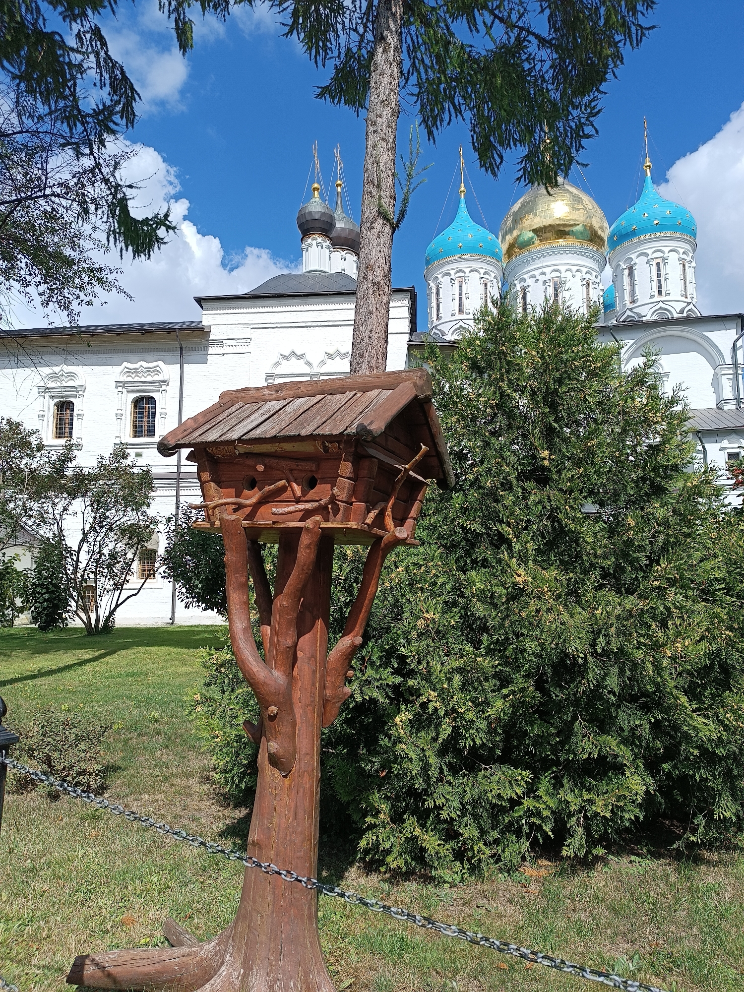 A wooden birdhouse, crafted to resemble a tree stump with branches, stands prominently in the foreground of an outdoor scene. The birdhouse is elevated on a weathered, wooden base that mimics roots. Behind the birdhouse, a large evergreen shrub dominates the mid-ground. In the background, a white building with arched windows and multiple domes, some gilded and some blue with gold stars, suggests a church or monastery. The sky above is a vibrant blue with scattered white clouds. The scene is set in a grassy area, with a chain barrier visible in the lower left corner.