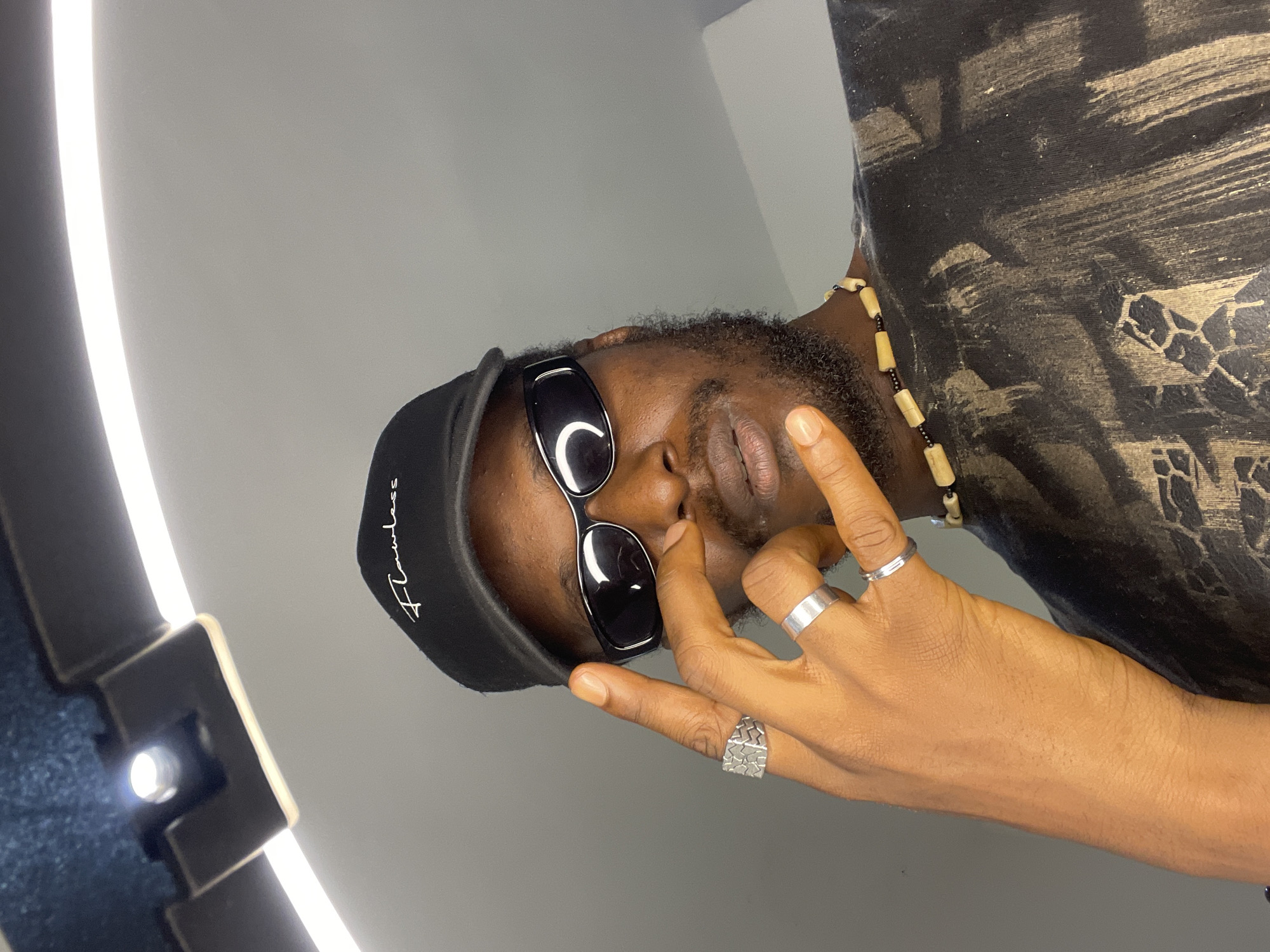 The image is a close-up, waist-up selfie of a Black man wearing a black baseball cap with "Flawless" embroidered in white, and black wraparound sunglasses. He has a beard and is wearing a black t-shirt with a distressed, abstract print in muted gold and black. A beaded necklace with tan cylindrical beads and small black beads is visible around his neck. His left hand is raised, with his index and pinky fingers extended in a gesture. He is wearing two silver rings on his left hand: a chunky, patterned signet ring on his index finger and a plain band on his ring finger. A third, thinner silver band is visible on his ring finger. The background is a plain, light gray wall. A bright, curved light fixture is visible at the top of the frame, casting a soft glow. There is no indication of the image being located in Abuja, Nigeria, or any other specific geographic location. The image appears to be taken indoors, likely in a personal space or studio setting.
