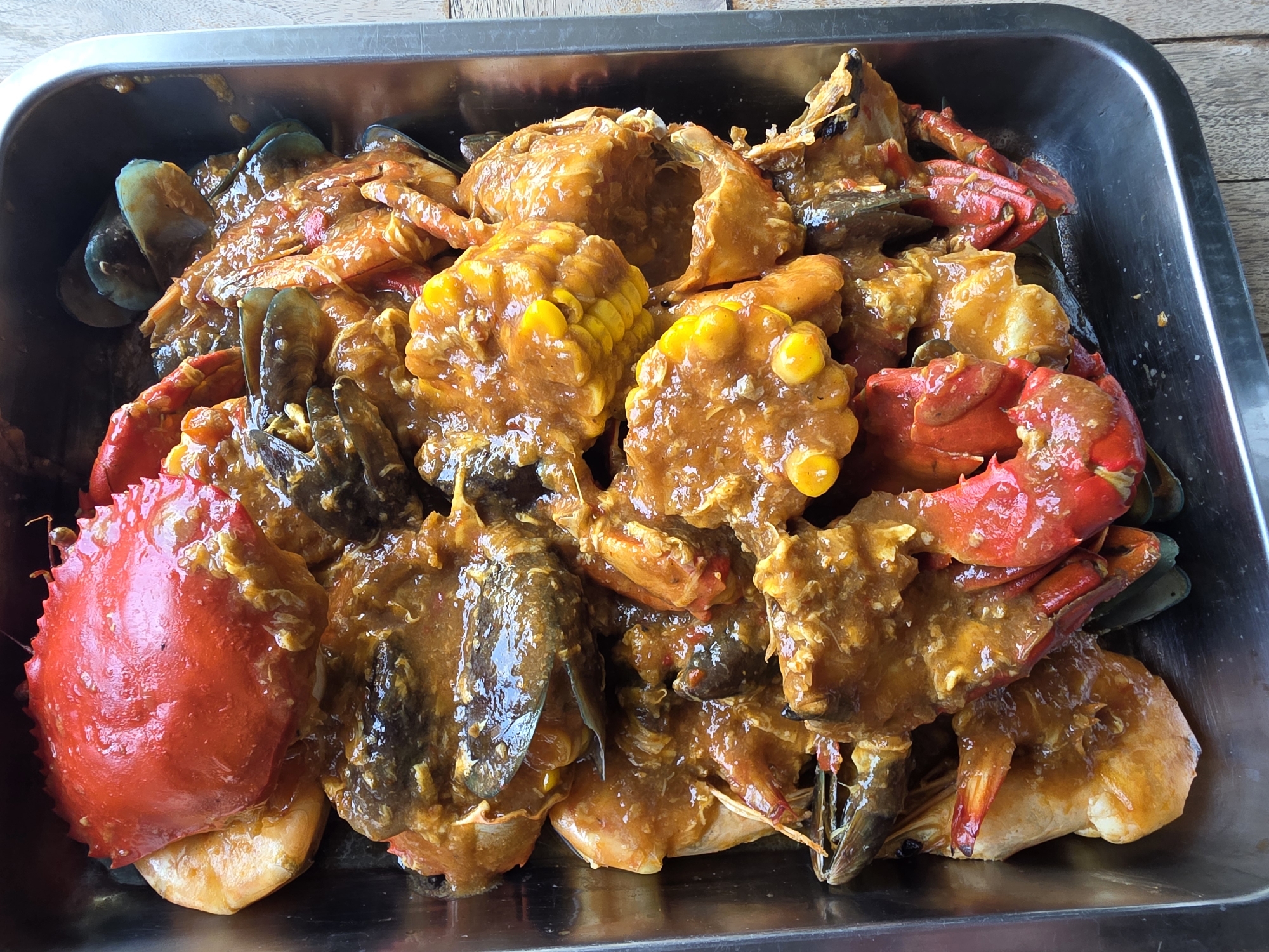 A close-up, overhead shot reveals a stainless steel rectangular pan overflowing with a seafood dish. The pan rests on a wooden surface. The contents of the pan are predominantly seafood coated in a thick, orange-brown sauce. Prominent items include whole red crabs, their shells glossy and iridescent. Mussels with dark blue-green shells are visible, partially submerged in the sauce. Several ears of corn, cut into sections, are scattered throughout the dish. The sauce appears to be a rich, savory mixture, possibly chili crab sauce or a similar Indonesian seafood preparation, clinging to the seafood and corn. The overall impression is one of abundance and a freshly prepared meal. This dish is likely found in a restaurant or food stall in Buleleng, Bali, Indonesia, given its presentation and common Indonesian ingredients.