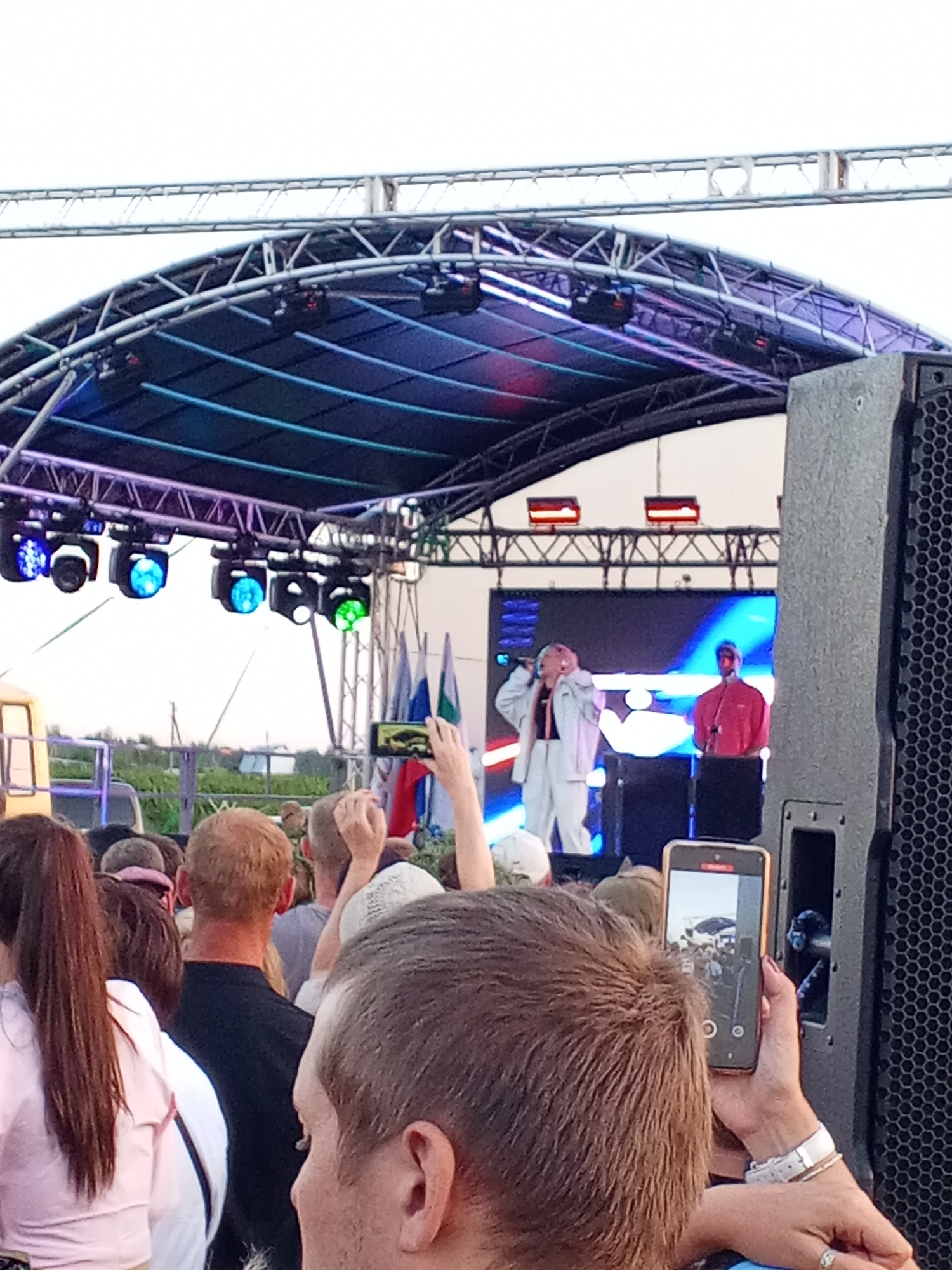 An outdoor concert is in progress, with a stage set up under a large, arched truss structure. The stage features a large LED screen displaying abstract blue and white patterns. Two performers are visible on stage: one in a white outfit with pink hair, holding a microphone, and another in a red shirt. Lighting rigs with blue and green lights are suspended from the truss. In the foreground, a crowd of people is gathered, some holding up their phones to record. One person in the foreground is holding a smartphone, capturing a video of the event. The environment appears to be a field or open area, with some greenery visible in the background and flags on the stage.