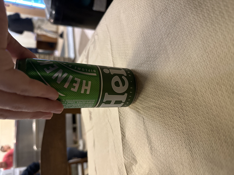 A hand holds a green Heineken beer can horizontally in a casual indoor setting in Nea Erithrea, Greece. The can prominently displays "HEINEKEN" in white capital letters. Other visible text includes "DIPLOME D'HONNEUR", "PREMIUM QUALITY", and "330ML". Below the volume, Cyrillic text "МПИРА-ΠΕΡΕΧ 330ml" is also present, indicating beer and its volume for a specific market. The hand gently grips the can.
The background is largely out of focus. A light-colored, textured surface, possibly a sofa arm or tablecloth, covers the right side of the image. In the blurred distance, indistinct figures of at least two people and various pieces of furniture are visible. One person on the left appears to be wearing a red top, and another further back has a blue garment. The scene suggests a relaxed atmosphere, with a person holding a beverage, likely in a home environment. The time of day is not discernible from the indoor lighting.