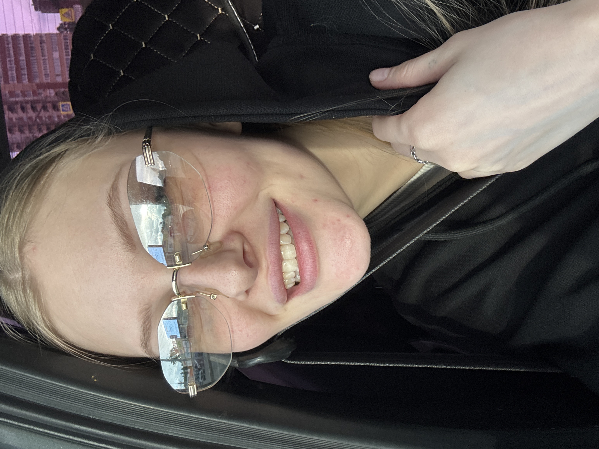A woman with light skin, wearing rimless glasses, is photographed from a close, slightly upward angle. She is smiling, revealing her teeth. She is wearing a black hooded garment, and her hand is visible, holding a part of the hood, with a ring on her finger. The reflection in her glasses shows buildings, indicating an urban environment. The location is Perm, Russia, as suggested by the urban landscape. The woman appears to be inside a vehicle, based on the dark, textured material visible at the bottom of the frame.