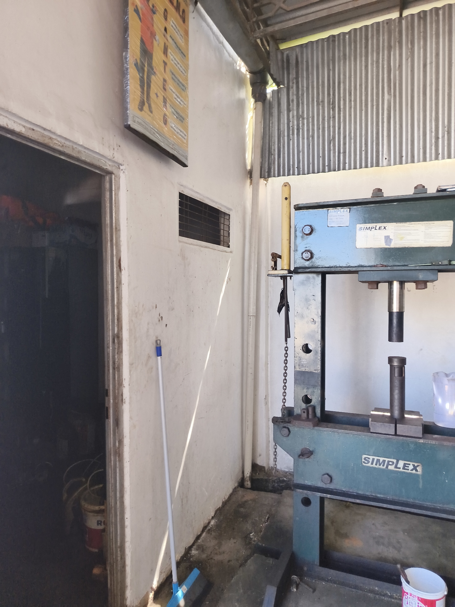 The image depicts an indoor workshop setting, likely in Pangkalpinang, Indonesia, characterized by a combination of industrial equipment and basic building materials. In the foreground and right of the frame, a large, green hydraulic press, identified by "SIMPLEX" branding, is prominently displayed. The press has a heavy-duty construction, with a substantial metal frame and a visible hydraulic cylinder. Resting on a shelf above the press is a white plastic container, possibly containing liquid.
To the left of the press, a white wall with a dirty texture forms the backdrop. A doorway is visible, leading into a dark interior where several buckets are stacked, one of which has the label "R" visible. A broom with a blue base and a silver handle stands upright against the wall near the doorway.
Further to the left, a yellow and red poster is affixed to the wall, encased in plastic. The poster contains text and images, though the details are partially obscured. A small, rectangular vent with a grid pattern is set into the wall above the poster.
The ceiling area shows exposed corrugated metal sheeting, suggesting a utilitarian construction. The overall environment appears to be a working area, with a practical and somewhat unkempt aesthetic, consistent with a small workshop or repair facility. Natural light enters from the right side of the image, casting shadows across the floor and wall.